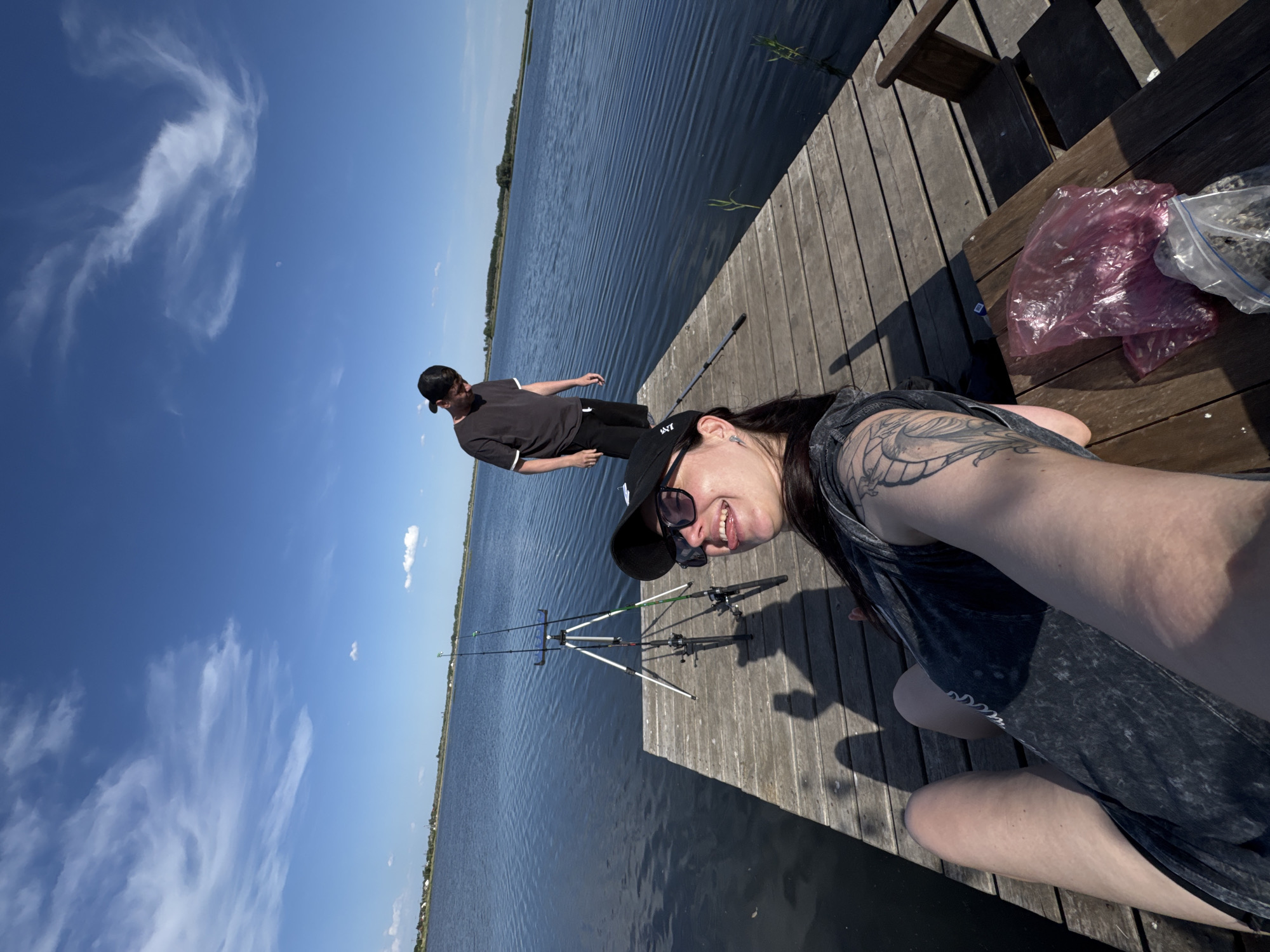 The image depicts a sunny outdoor scene on a wooden dock. A woman wearing sunglasses and a baseball cap is in the foreground, taking a selfie. She has a visible tattoo on her arm. In the background, a man in a similar cap and dark clothing is walking along the dock towards the water. Two fishing poles on a stand are positioned near the edge of the dock. A table with some bags sits on the right side of the dock. The location appears to be a lake or large body of water, with land visible on the opposite shore. The sky is blue with scattered clouds. The city is Unknown, Unknown.