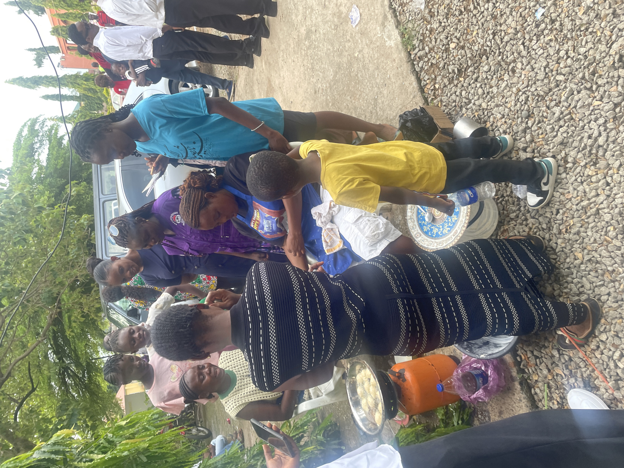 In the city of Gwagwalada, Nigeria, a group of people are gathered outdoors, possibly for a community event or a marketplace. The scene is set on a paved area with gravel surrounding it, under the shade of trees. In the foreground, a woman in a dark patterned dress is standing over a gas cylinder and a frying pan filled with what appears to be food being fried. Beside her, a young boy in a yellow t-shirt and dark pants is observing the cooking. Several other individuals, mostly women and girls, are clustered around, watching the process or engaged in conversation. Some are dressed in casual clothing, while others wear more formal attire, including white shirts and dark pants, suggesting a mix of participants. A vehicle, possibly a van, is visible in the background, along with other people walking or standing. The overall atmosphere seems to be one of social interaction and food preparation.
