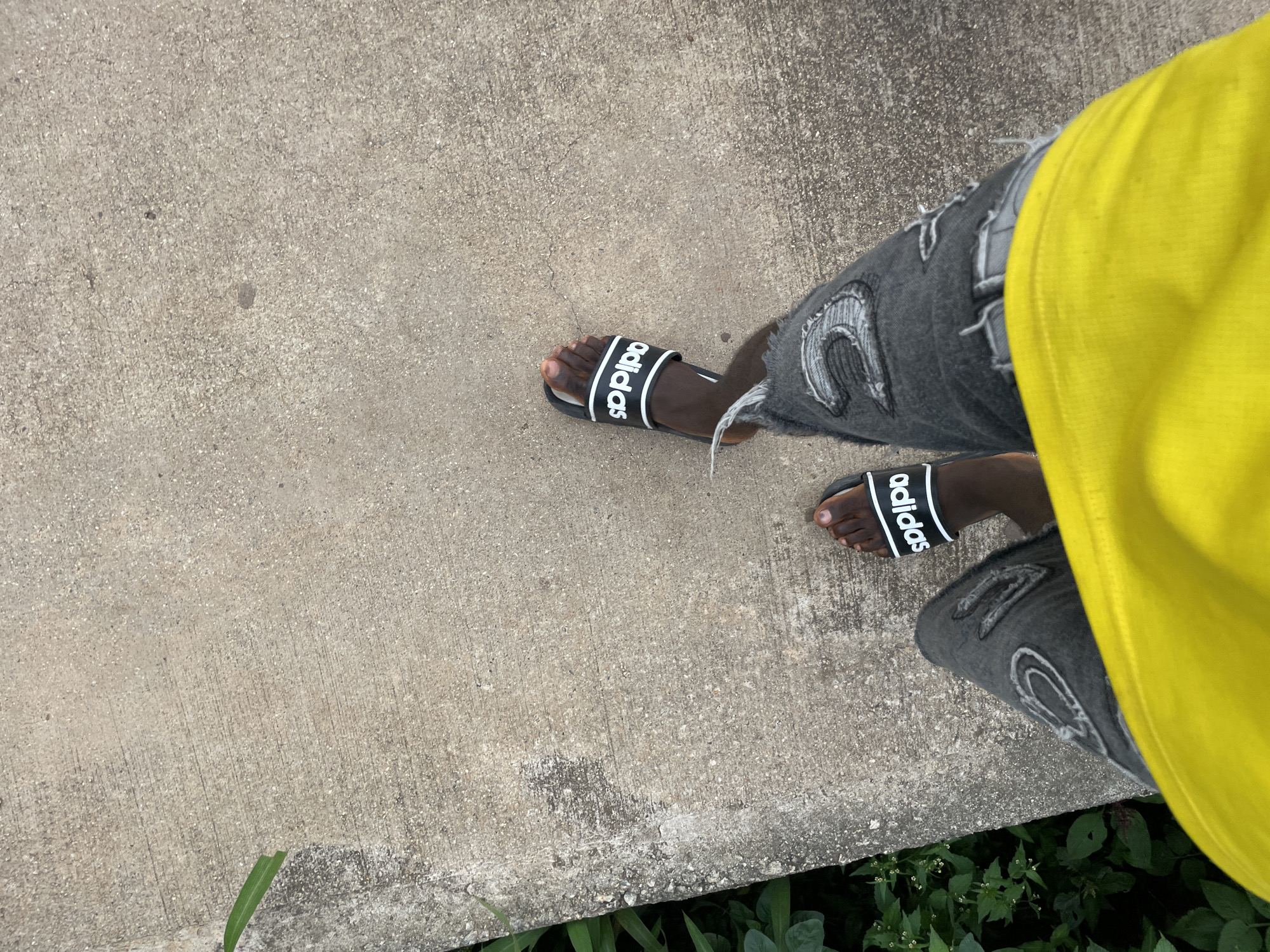 The image shows a person's legs from the knees down, standing on a concrete surface. The person is wearing ripped black denim jeans and black Adidas slides. The left leg is partially visible with the right leg more prominently displayed, showing the Adidas branding on the slide. To the left of the concrete surface, there is lush green foliage. The concrete appears to be outdoors, possibly a sidewalk or a pathway in the city of Jos, Nigeria. The lighting suggests it is daytime.