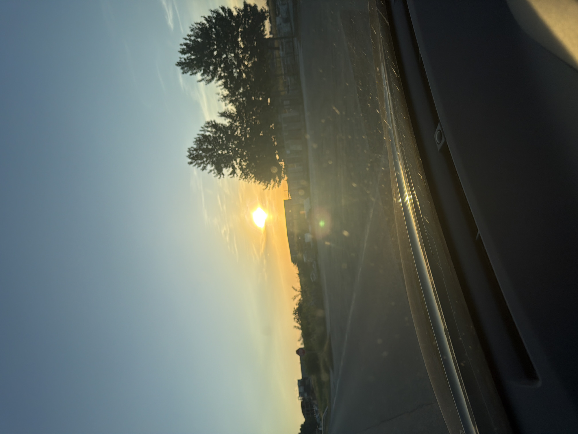 The image appears to be taken from inside a vehicle. A bright sun is partially obscured by trees and buildings in the distance. The sky is a gradient of blue to orange near the horizon. Part of the vehicle's interior is visible in the foreground. The location is not identifiable as Branipole, Bulgaria from this image alone. There is no evidence of people, specific objects, or actions discernible.