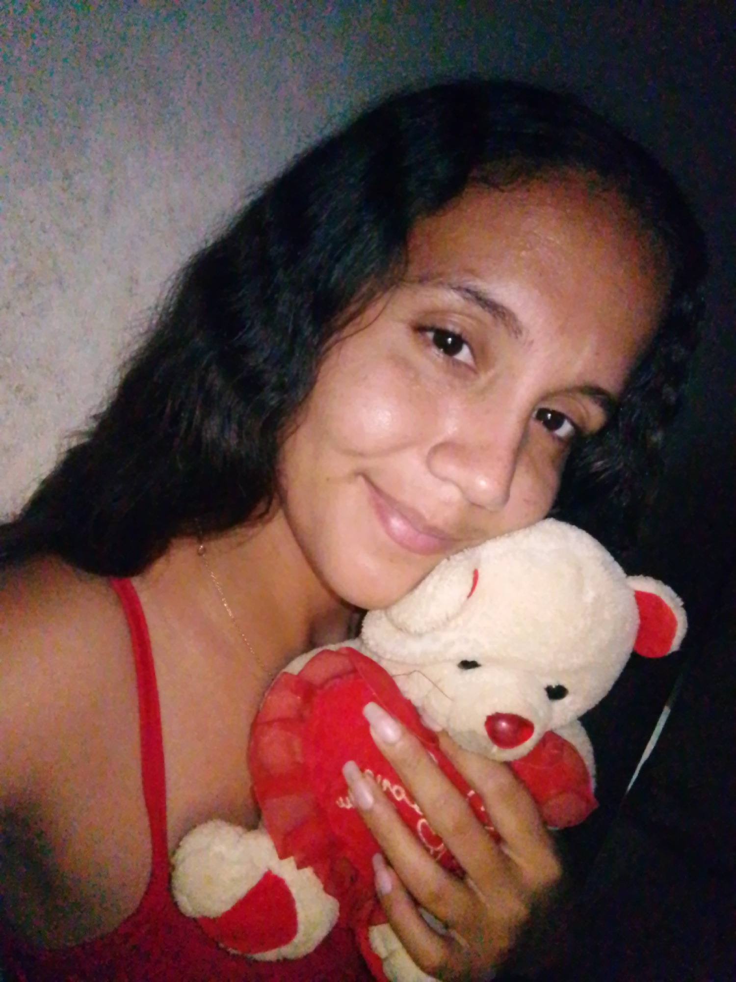 A woman with dark hair and skin is holding a stuffed bear. She is wearing a red top. The background appears to be a wall or dim interior space. The location information is not discernible from the image.