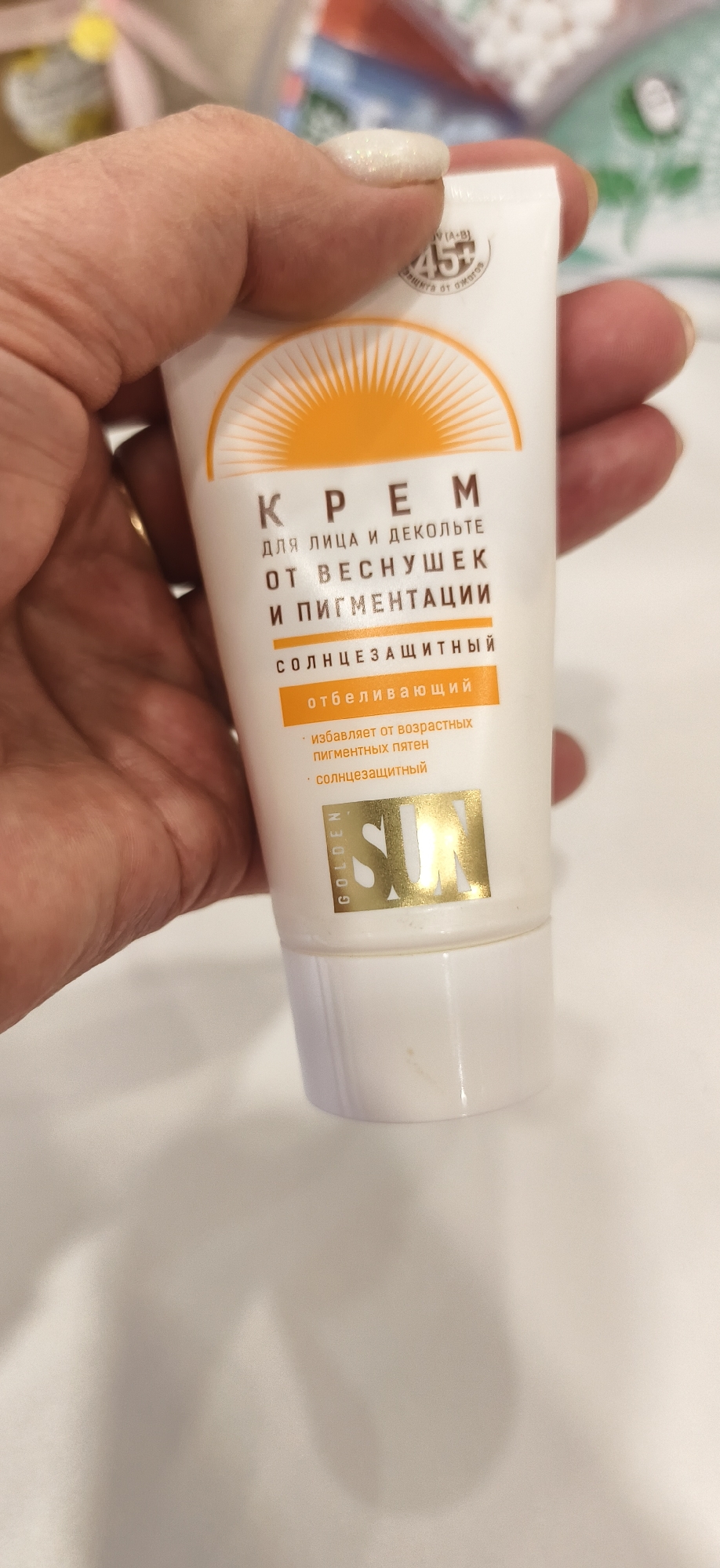 A photograph depicts a hand holding a tube of cosmetic cream. The product is labelled in Russian and features the words "Крем для лица и декольте от веснушек и пигментации" (Cream for face and décolleté from freckles and pigmentation). The tube also states "солнцезащитный" (sunscreen). The product claims to lighten age spots and provide sun protection. The brand "GOLDEN SUN" is also visible on the tube. The background is slightly blurred and out of focus.