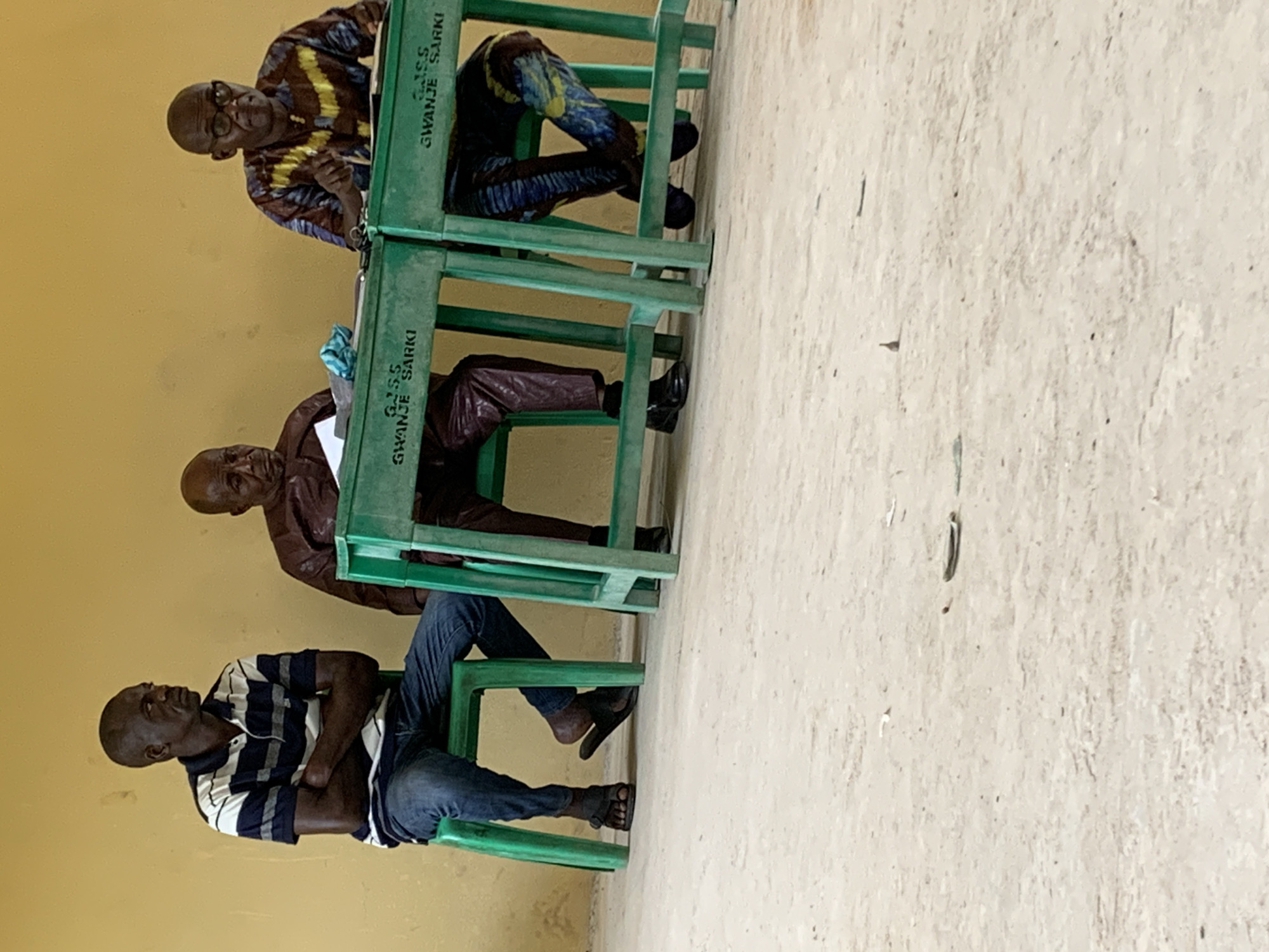 Three men are sitting on green plastic chairs in what appears to be an indoor setting, possibly a waiting area or office in Akwanga, Nigeria. The men are seated in a vertical stack from the viewer's perspective. The man at the bottom is wearing a striped shirt and jeans, and sandals. The middle man wears a brown jacket and pants, and black shoes. The man on top is wearing a colorful patterned shirt and pants, and glasses. All three men are bald. The chairs are labeled "G.S.S GWANJE SARKI". The background consists of a yellow wall and a light gray floor. The image is slightly tilted.
