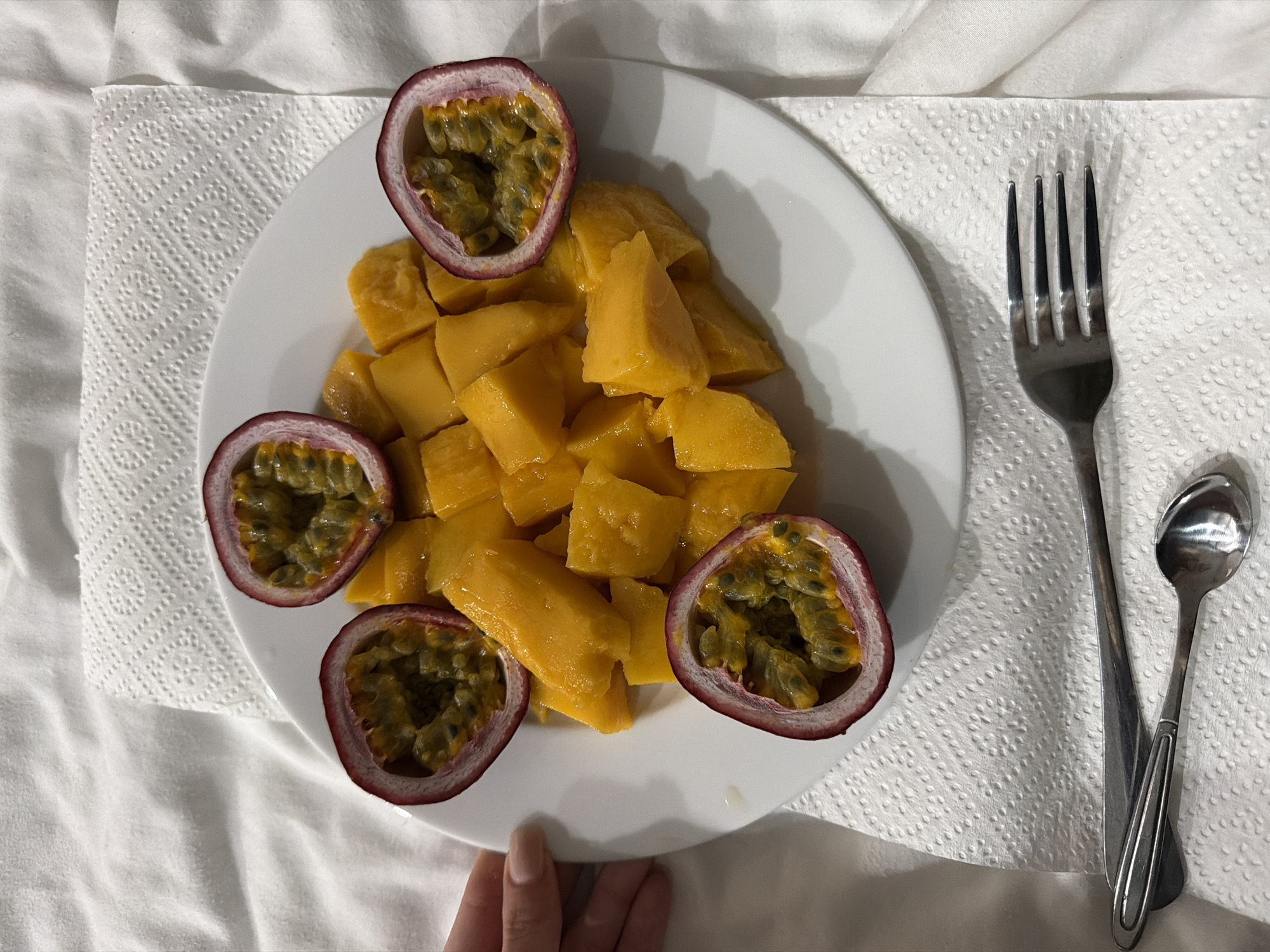 A white plate containing diced mango and three halved passion fruits sits on a white surface with a fork and spoon to the side. The background appears to be a white, textured cloth. A hand is visible at the bottom left corner of the image. The exact location in the city of Đà Nẵng, Vietnam cannot be determined from the provided image alone.