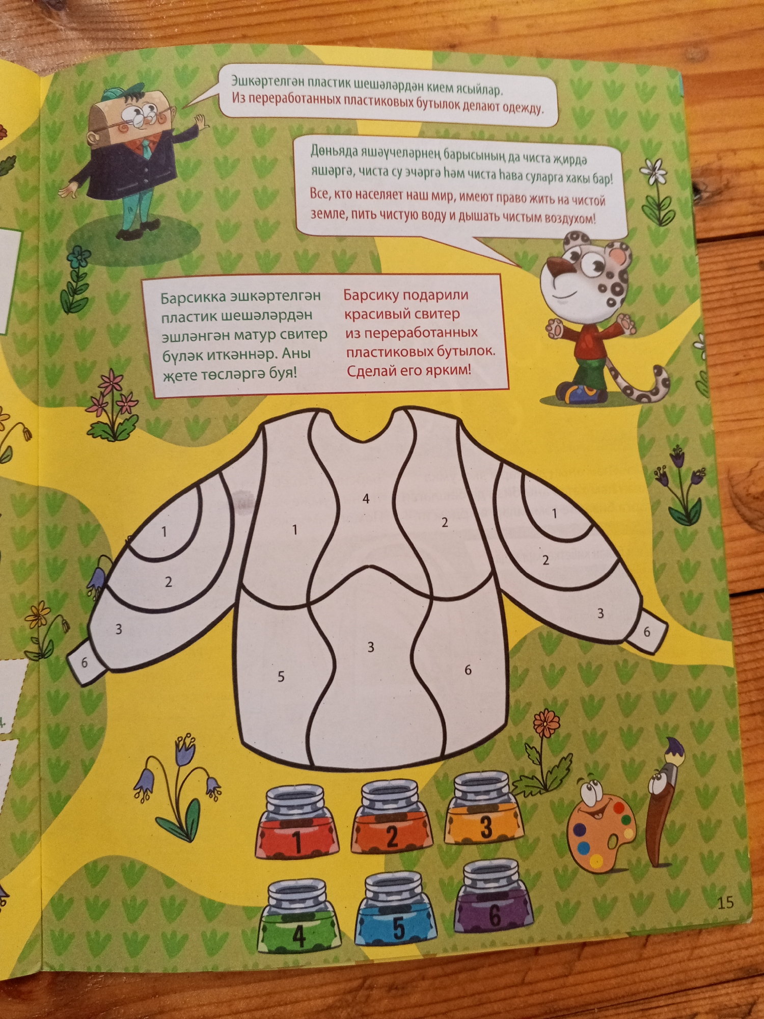 The image shows a page from a children's activity book featuring a coloring section with a sweater outline and color palettes. The page is set against a wooden table surface, suggesting it's a physical book being viewed. There are no specific geographical indicators, including the city of Chistopol, Russia, present in the image. The content includes illustrations of a character made of a cardboard box and a leopard-like creature, along with text in a language that appears to be Tatar or a Turkic language, and Russian. The activity involves coloring sections of a sweater outline based on numbered color choices. The color palettes consist of jars labeled with numbers 1 through 6, representing different colors. The overall environment depicted is a children's activity page, not a real-world scene in Chistopol or elsewhere.