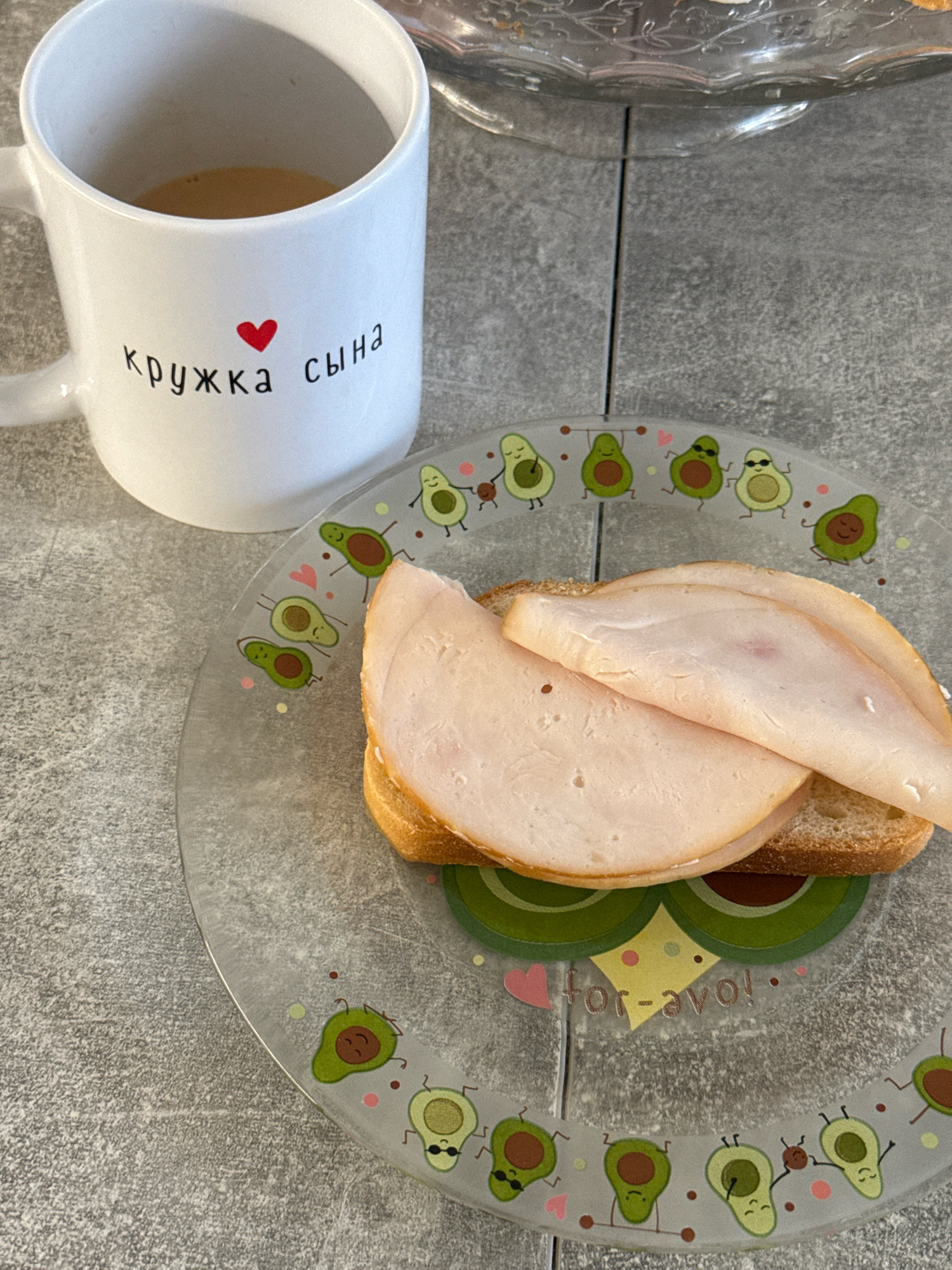 The image displays a close-up of a meal on a grey, textured surface, possibly a countertop or table. A white mug with a red heart and Russian text reading "кружка сына" (mug for son) is visible on the left. Inside the mug, there is a light brown liquid, likely coffee or tea. To the right of the mug, a transparent glass plate with an avocado-themed design is present. The plate holds a sandwich consisting of two slices of bread topped with several slices of processed meat, possibly ham or turkey. The plate's design features cartoon avocados in various poses, some with sunglasses, and the text "for-avo!". The scene is lit from above, casting subtle shadows. There is no visible indication of the city of Lyubertsy, Russia, or any specific outdoor environmental details. The image appears to be a still-life composition focusing on breakfast or a snack.