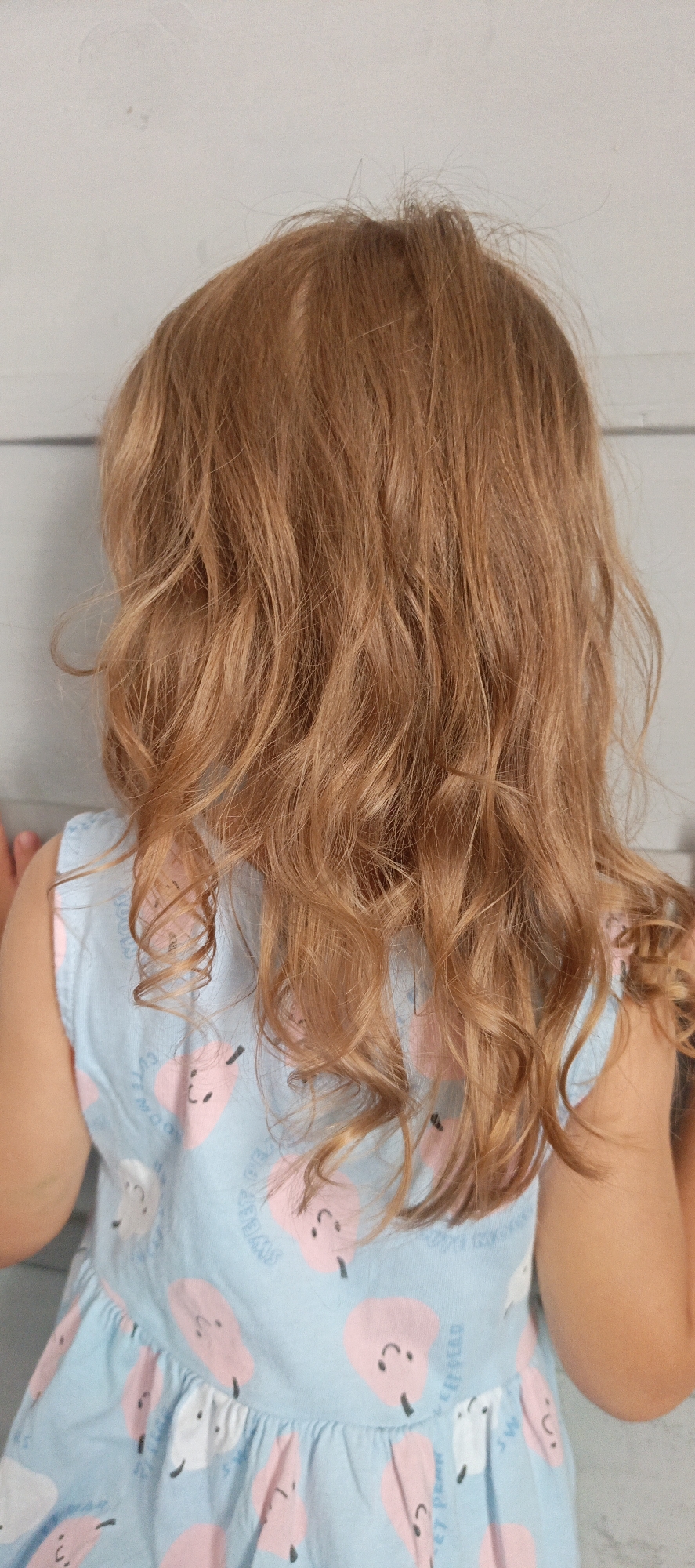 This image, taken outdoors in Gorno-Altaysk, Russia, shows the back of a young child with reddish-blonde, wavy hair. The child is wearing a light blue sleeveless dress adorned with a pattern of smiling pink and white pears. The background consists of a light-colored, textured wall, possibly made of wood panels. The lighting is natural, suggesting it is daytime.