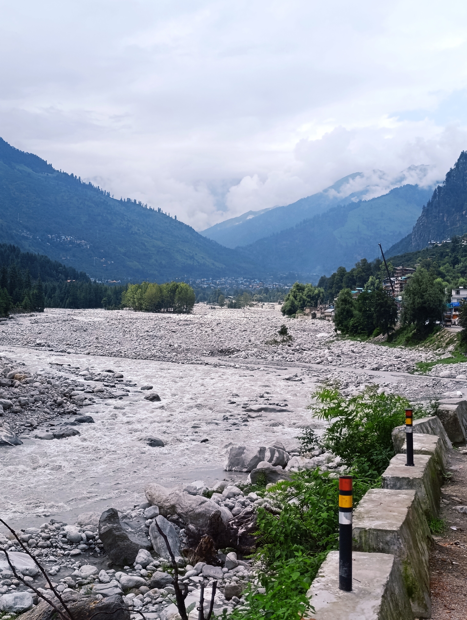 The image depicts a wide, rocky riverbed in the Vashisht region of India, with a turbulent river flowing through it. On the left side of the river, a dense forest of pine trees blankets a steep mountainside. In the distance, rolling hills and mountains are visible, shrouded in a light mist under a cloudy sky. The right bank of the river features a more developed area with buildings constructed on the hillside, interspersed with trees. A concrete retaining wall, marked with bollards, runs along the edge of a road adjacent to the river. The overall scene conveys a natural, mountainous landscape with signs of human settlement.