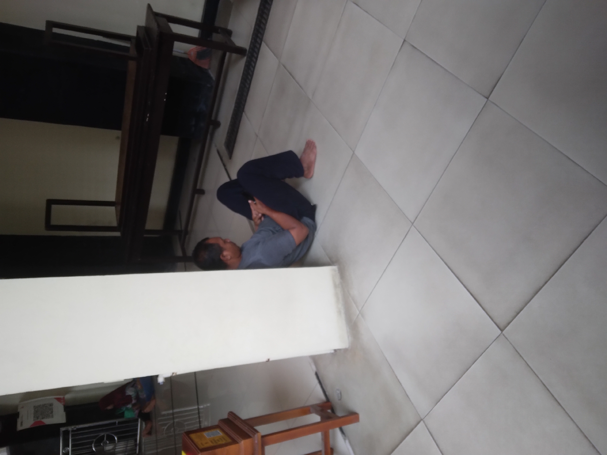 A man is sitting on the floor, leaning against a white pillar, and looking at his phone. He is wearing a gray t-shirt and dark pants, with his bare feet visible. The floor is tiled with large, square, light-colored tiles arranged diagonally. To the left of the man is a wooden stand with a framed object on it. In the background, there is a long, dark wooden table with two upright supports, and a person's legs are visible underneath it. Further back, there is a dark wall and a metal railing with a QR code poster attached to it. The environment appears to be an indoor public space.