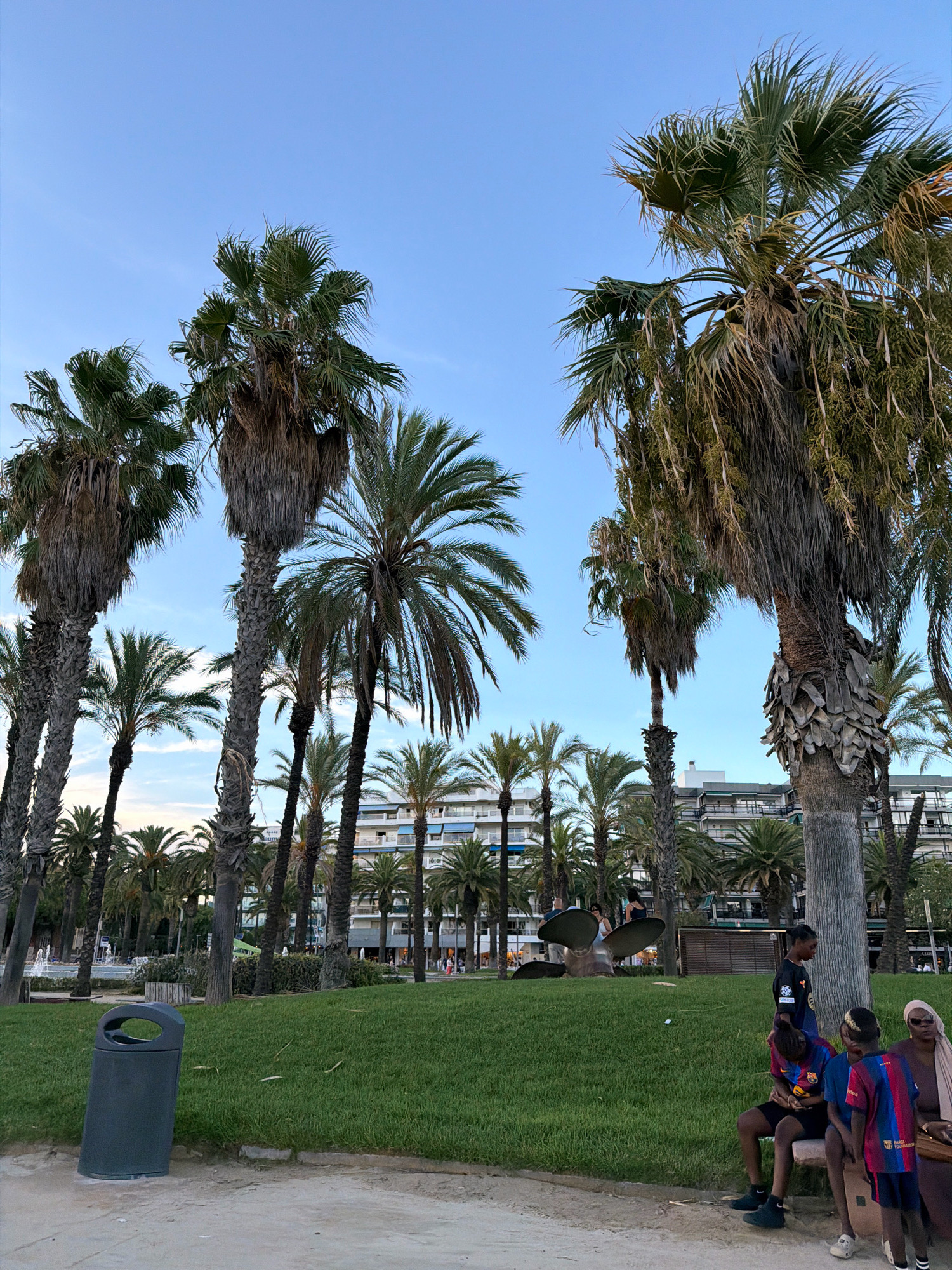 The image depicts a public area in Salou, Spain, characterized by a profusion of palm trees. In the foreground, a dark gray trash receptacle stands on a paved surface adjacent to a manicured lawn. Several individuals are visible, some seated on a bench and others standing. A group of young people, one wearing a dark blue shirt with orange lettering and another in a Barcelona-themed jersey, are interacting. Further in the background, a large metallic sculpture resembling a propeller is situated on the grassy area. Behind the palm trees, a multi-story building with balconies and blue awnings is visible, suggesting a coastal urban setting. The sky is clear and blue, indicating daytime.