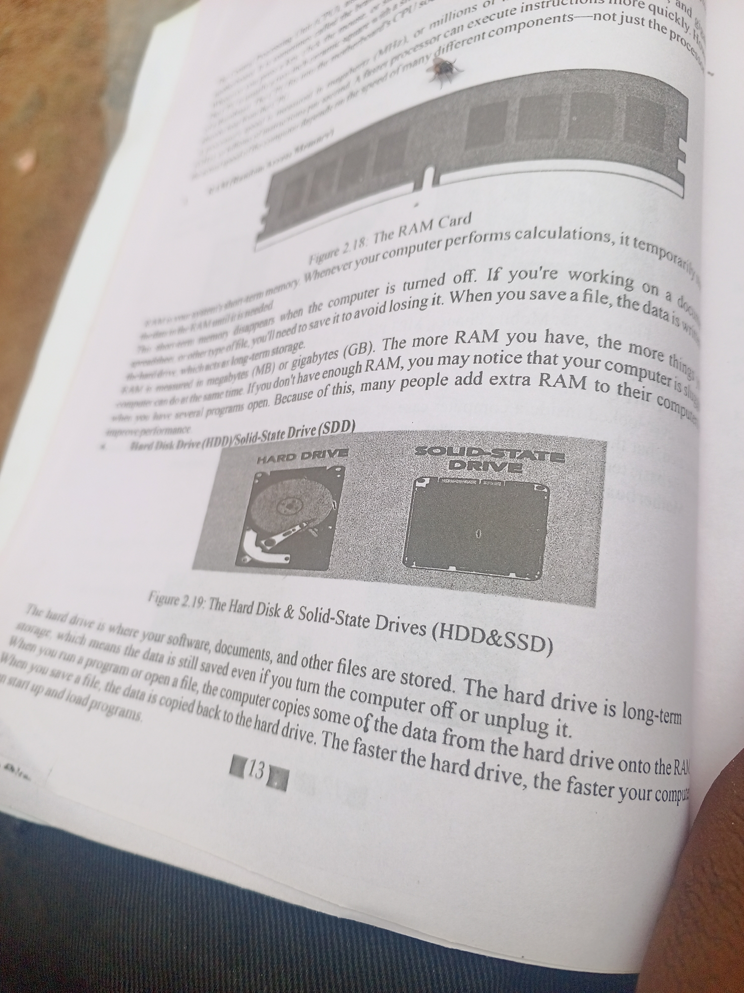 This is a close-up photograph of an open book, taken from a slightly high angle. The page is filled with text and two illustrations. The subject matter appears to be educational, specifically related to computer hardware.
The first illustration, at the top of the page, shows a close-up of a RAM stick, a common computer component. Below this, a larger illustration is divided into two halves. The left half shows an image labeled "HARD DRIVE" which depicts the internal components of a traditional hard disk drive, including a spinning platter and read/write heads. The right half shows a flat, rectangular object labeled "SOLID-STATE DRIVE", which is a more modern type of storage device.
The text on the page explains the functions of RAM, hard disk drives, and solid-state drives. It discusses how RAM is temporary memory and how hard drives and SSDs provide long-term storage. Some of the text is also visible on the pages surrounding the main subject, suggesting it is part of a larger book. There is a number "13" visible at the bottom of the page, likely a page number.
There are no people or specific location cues within the image itself, other than what can be inferred from the educational content. The lighting appears to be artificial, and the setting is indoors, as indicated by the book and the overall presentation. No specific time of day or weather conditions are discernible. The overall mood is informative and academic.