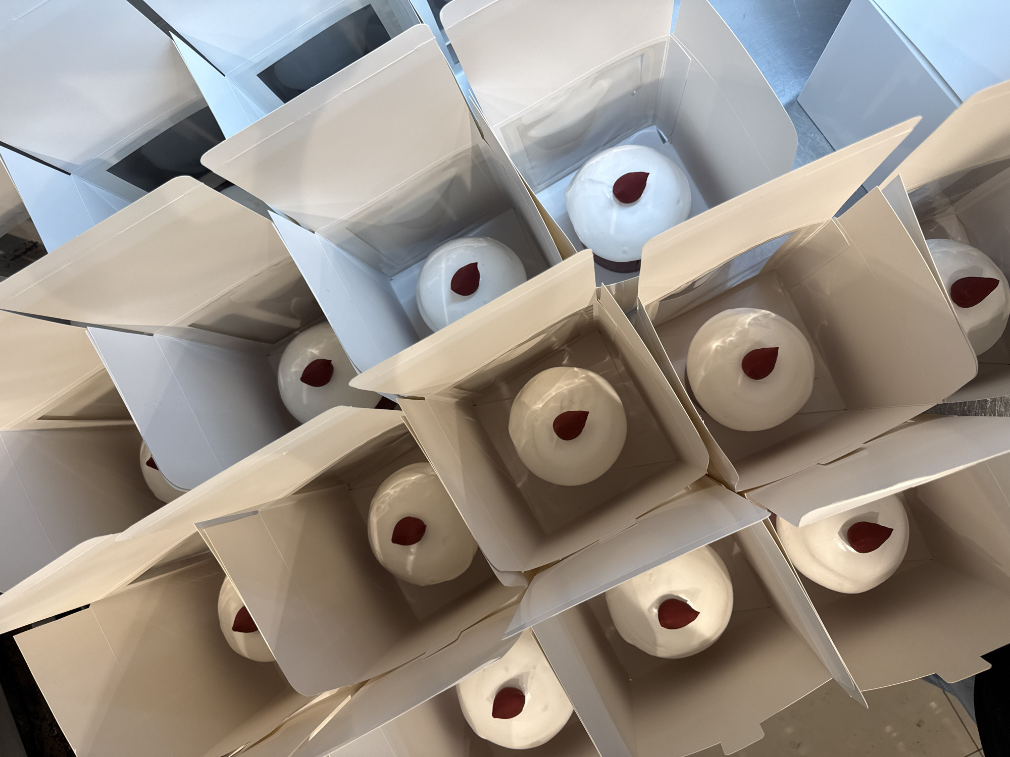 The image shows a collection of white, square pastry boxes. Each open box contains a single, round dessert, likely a cake or pastry, covered in white icing with a red, droplet-shaped decoration on top. The arrangement appears to be in a commercial or catering setting, possibly in Belgrade, Serbia, but without further context, this location cannot be definitively confirmed. There are no people visible in the image, and the focus is solely on the pastries and their packaging.