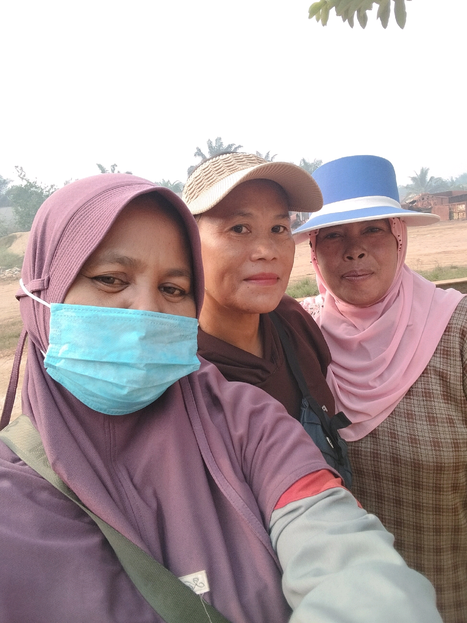 The image shows three women posing for a selfie outdoors. The woman in the foreground, on the left, is wearing a purple hijab and a light blue surgical mask. She has a green strap across her chest, likely from a bag. To her right, a second woman is wearing a tan baseball cap and a brown top. The third woman, on the far right, is wearing a light blue wide-brimmed hat and a pink hijab. She has a brown plaid shirt underneath. The background suggests a rural or semi-urban environment with some trees and what appears to be industrial structures or construction in the distance. The lighting indicates it is daytime, with the sky being bright and overcast. The location is specified as Teluk Tenggirik, Indonesia.
