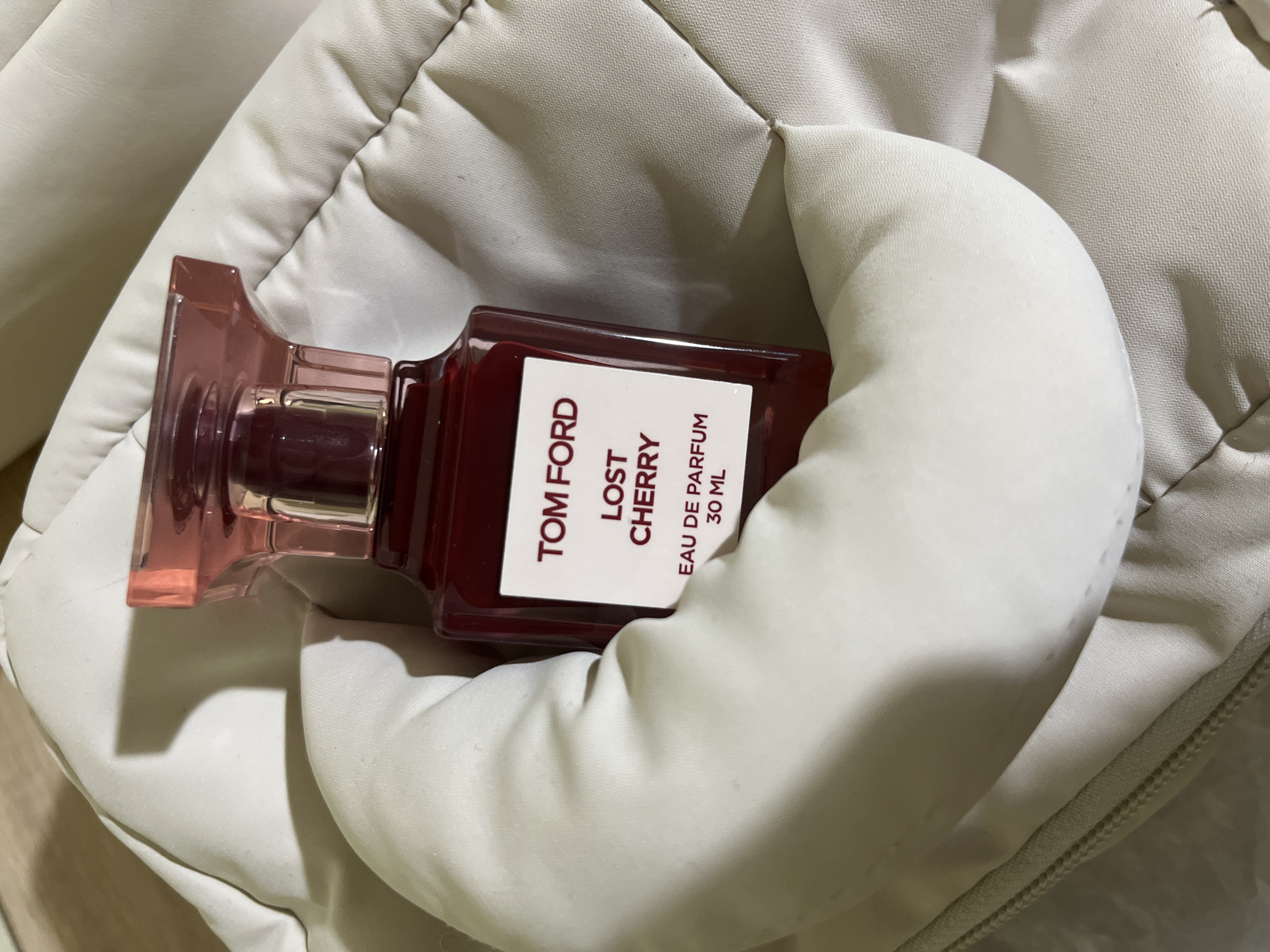 A Tom Ford Lost Cherry Eau de Parfum 30ml bottle rests nestled within a cream-colored, quilted fabric. The bottle is a deep red color, with a clear pink cap. The label on the bottle is white with red text. The fabric appears to be soft and plush, with visible stitching and a zipper along the bottom edge. The lighting suggests an indoor setting, with shadows cast on the fabric. There are no people or other objects visible in the image. The location is Unknown, Unknown.