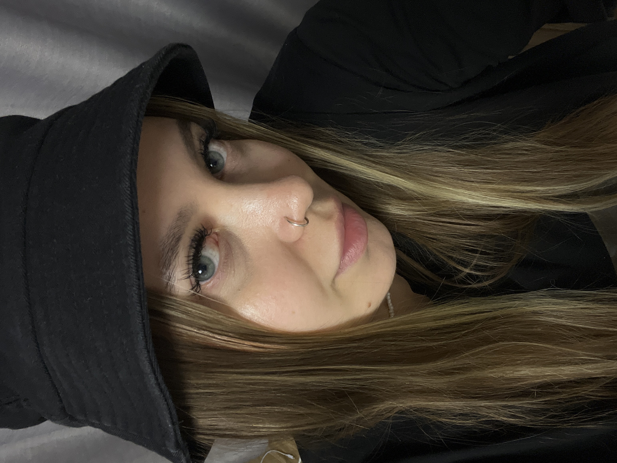 This is a close-up, vertically oriented portrait of a young woman taken indoors. Her head is tilted, giving the image a candid and intimate feel.
The primary subject is a woman with fair skin, light blue-grey eyes, and long, straight to slightly wavy hair that features blonde and light brown highlights. Her left eye is prominently displayed, framed by long, dark eyelashes. She has a small silver hoop nose ring adorning her septum and her lips are softly parted, showing a natural pink color. A subtle mole is visible on her upper right cheek.
She is dressed in a black bucket hat, which covers part of her forehead, and a black garment, possibly a jacket or top, visible around her shoulders and neck. A delicate necklace, appearing to be made of small white or light-colored beads, is visible just below her chin.
The background is softly blurred, showing a muted grey texture that suggests an indoor environment, possibly a wall or fabric. The lighting is gentle and diffused, contributing to a quiet, natural mood. The woman's expression is neutral and calm as she looks towards the camera. There is no visible text in the image. The provided location context of Krasnodar, Russia, is not visually discernible within the photograph.