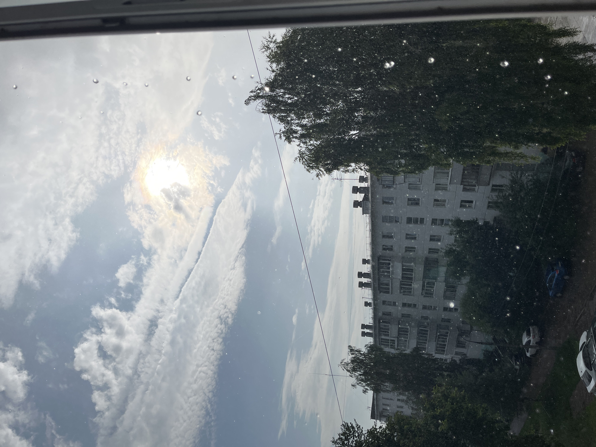 The image depicts an outdoor scene in Chaikovsky, Russia, viewed through a window with water droplets on the glass. The sky is partly cloudy with the sun visible, creating a rainbow effect. Below the sky is a multi-story residential building with a light-colored facade. Trees, including a large one with drooping branches in the foreground, are situated in front of and around the building. Several cars are parked on the street in front of the building, and what appears to be rain is falling, as indicated by the droplets on the window and scattered throughout the scene.