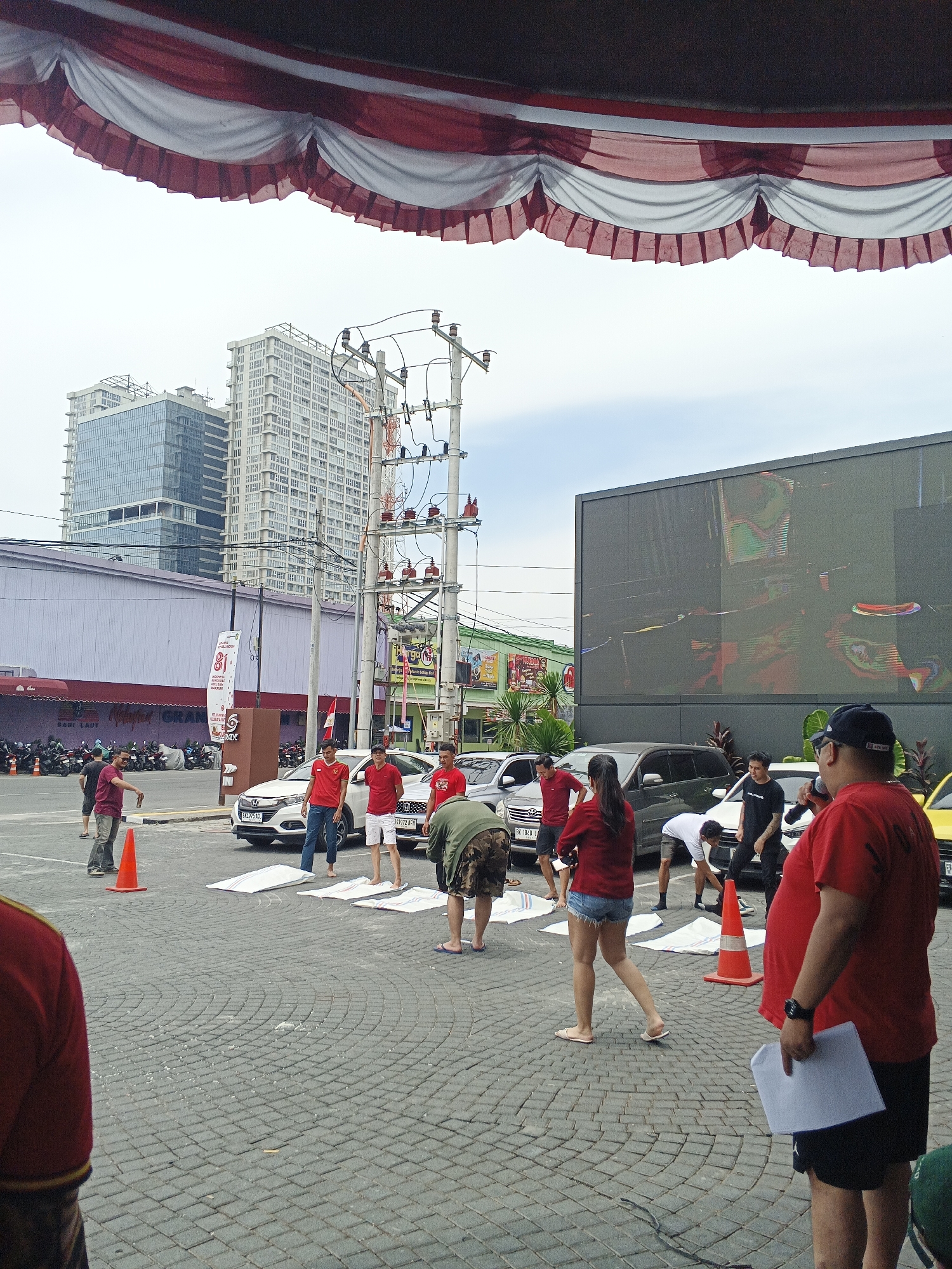 In front of the Palladium Mall in Medan, Indonesia, a group of people are gathered in an outdoor plaza. The plaza is paved with interlocking grey bricks. Several cars are parked in the background. A large digital screen is mounted on the side of a building, displaying abstract colorful patterns. Overhead, a red and white striped awning provides shade. Some individuals are wearing red shirts, and some are engaged in an activity involving white mats laid out on the ground. There is an orange traffic cone visible near the center of the image. The sky is overcast.