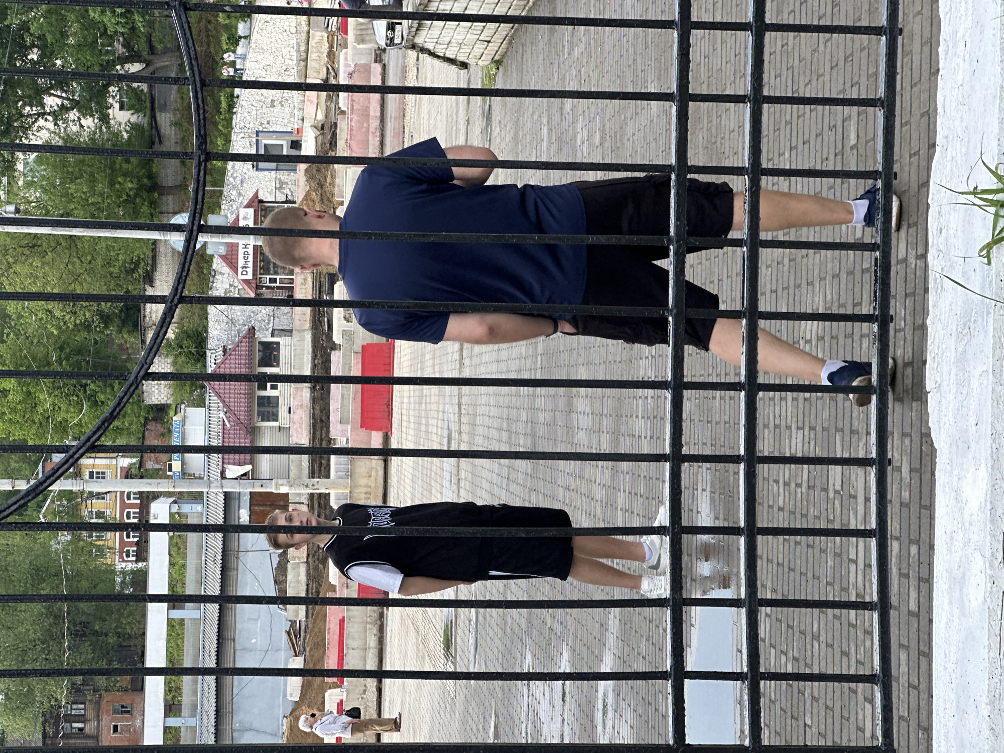 Observed from behind a black iron fence, a man in a navy blue t-shirt and black shorts stands above a second man wearing a black and white athletic jersey and black shorts. Both men are standing on a gray brick-paved area. In the background, buildings and trees can be seen. The street appears to be in Vladimir, Russia, and under construction or renovation.