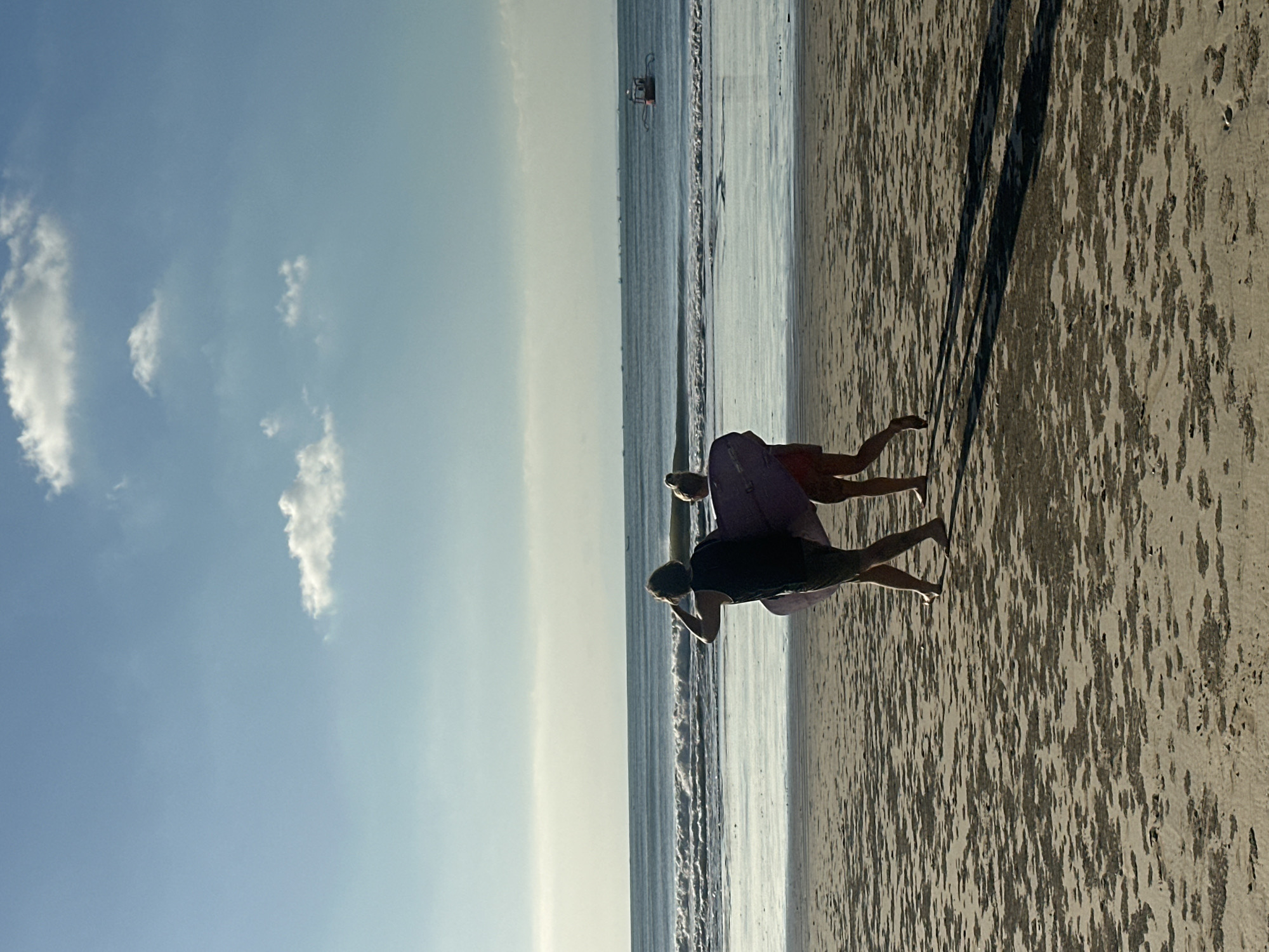 Two individuals, one male and one female, are walking away from the viewer on a sandy beach in Kuta Selatan, Indonesia. The male, on the left, is wearing a dark tank top and shorts. He is carrying a purple surfboard under his left arm and has his right hand near his head, shielding his eyes. The female, to his right, is wearing shorts and is carrying the same purple surfboard with him, its front end extending out to her right. The surf board is positioned between them, with the male holding the back and the female holding the front. They are walking towards the ocean, which is visible in the background, with gentle waves rolling onto the shore. A small, distant boat is visible on the horizon. The sky is blue with scattered white clouds, and the sun appears to be casting long shadows of the two people onto the sand. The beach is wet and has scattered patches of darker, damp sand.
