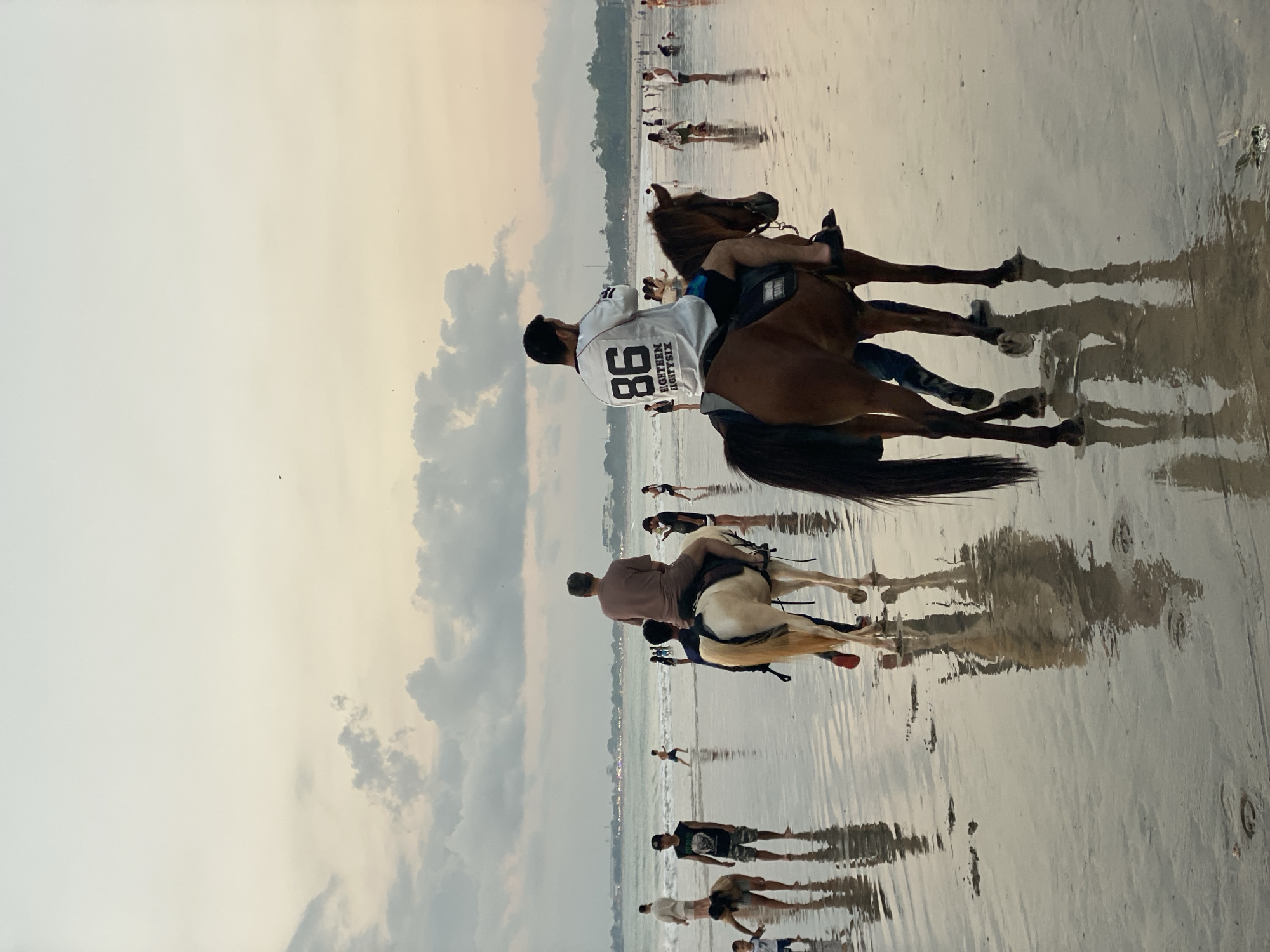 This image captures a tranquil late afternoon or early evening scene on a wide, wet sandy beach in Kuta Selatan, Indonesia. The sky is largely overcast with soft, diffuse light and scattered clouds, suggesting either an approaching sunset or an overall cloudy day.
In the foreground, two individuals are riding horses along the water's edge. The rider in the most prominent position wears a white long-sleeved shirt with the large number "86" and the text "EIGHTEEN EIGHTYSIX" printed vertically below it on the back. This rider is on a dark brown horse. To their left, a second rider is visible on a lighter-colored, possibly cream or palomino, horse. Both horses and riders are reflected with remarkable clarity on the highly reflective, mirror-like wet sand, indicating a very calm sea or low tide.
Further in the mid-ground and background, numerous other people are scattered across the beach, walking, standing, or enjoying the shoreline. The distant horizon reveals the ocean and a faint outline of the coastline, featuring a line of vegetation and some indistinct structures or lights, hinting at nearby development. The overall ambiance is leisurely and peaceful, with individuals enjoying recreational activities on the beach.