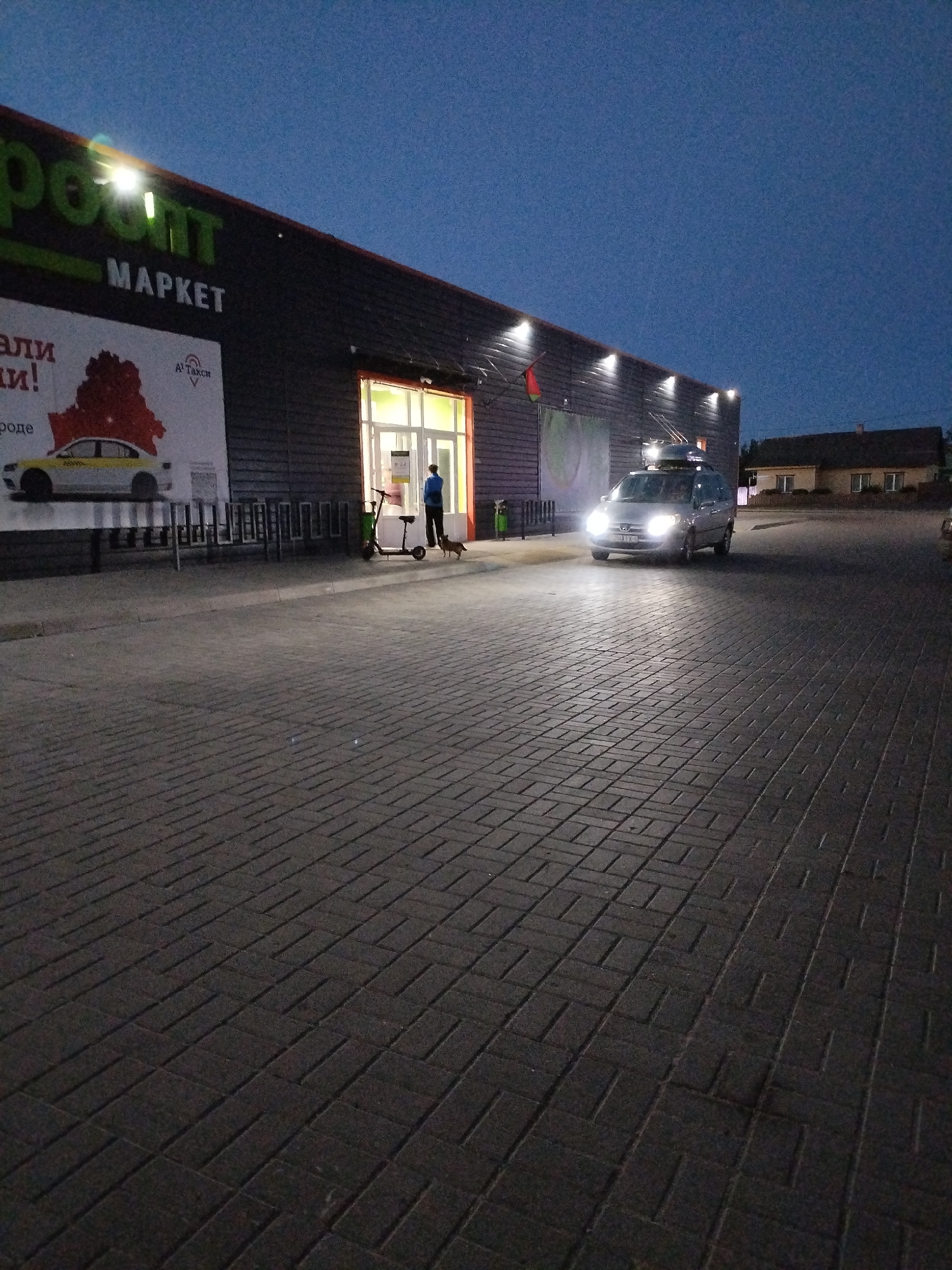 An outdoor, medium-range, eye-level shot captures the exterior of a "Pozit-Market" supermarket in Bialynichy, Belarus, during twilight. The building's dark, slatted facade is illuminated by overhead lights and light emanating from the brightly lit entrance. To the left, a large advertisement features a red outline of Belarus, a yellow taxi, and text in Cyrillic. A bicycle rack is situated in front of the building.
In front of the market entrance, a person in a blue jacket stands beside a green electric scooter and a small dog. A silver minivan with its headlights on is driving towards the viewer on a brick-paved area in front of the market, creating light reflections on the ground. A residential building with a dark roof and multiple windows is visible in the background on the right. The sky is a deep blue, indicating dusk.