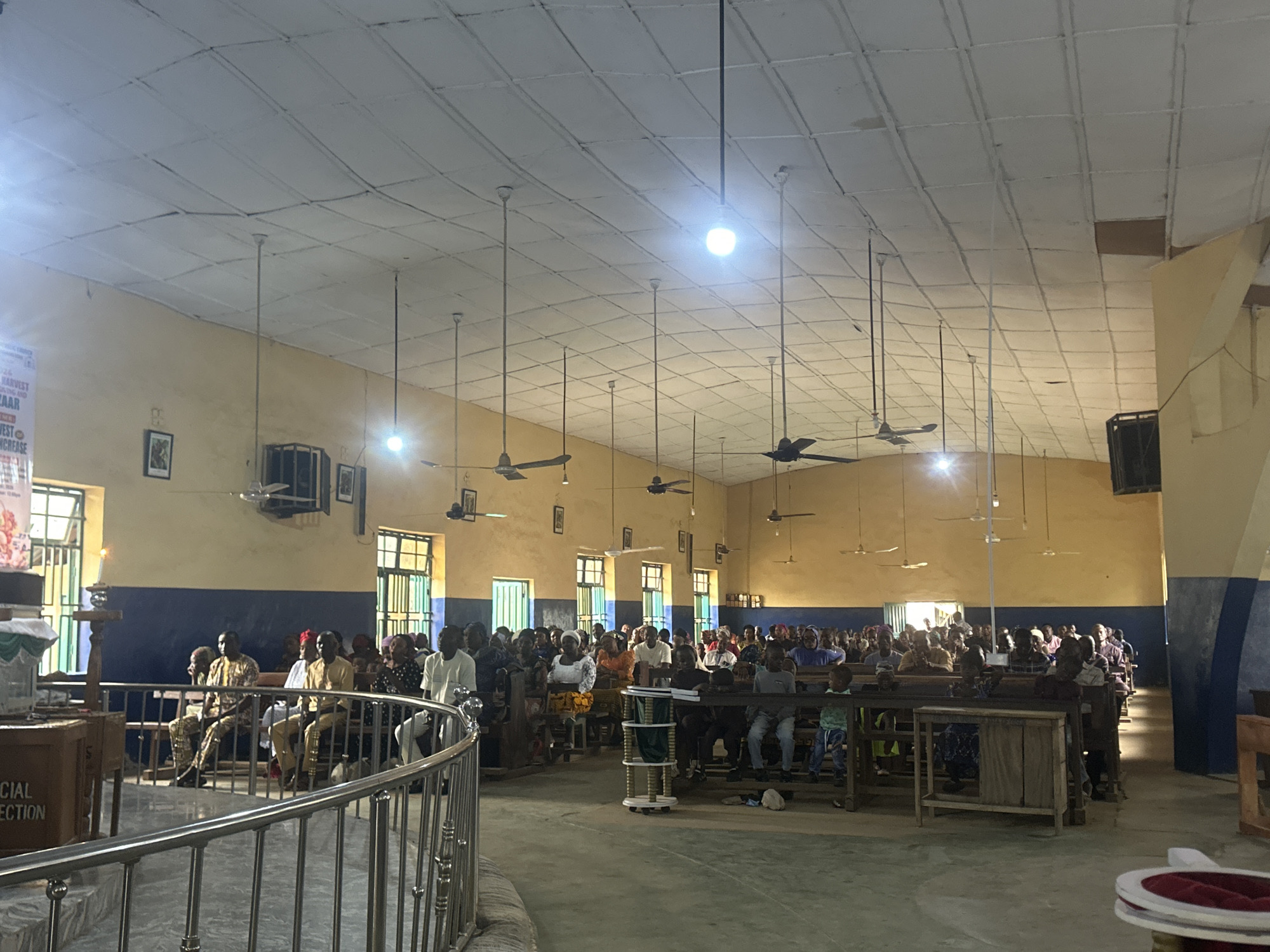 The image depicts the interior of a church in Akwanga, Nigeria, with a congregation seated in pews. The church has a pale yellow and blue painted interior with a white, tiled ceiling. Several ceiling fans are visible, along with hanging light fixtures, some of which are illuminated. Two framed pictures hang on the wall. In the foreground, a metal railing separates the viewer from the seating area. People of various ages are seated in the pews, some facing forward, others appearing to be in conversation. The atmosphere appears to be during a service or gathering.