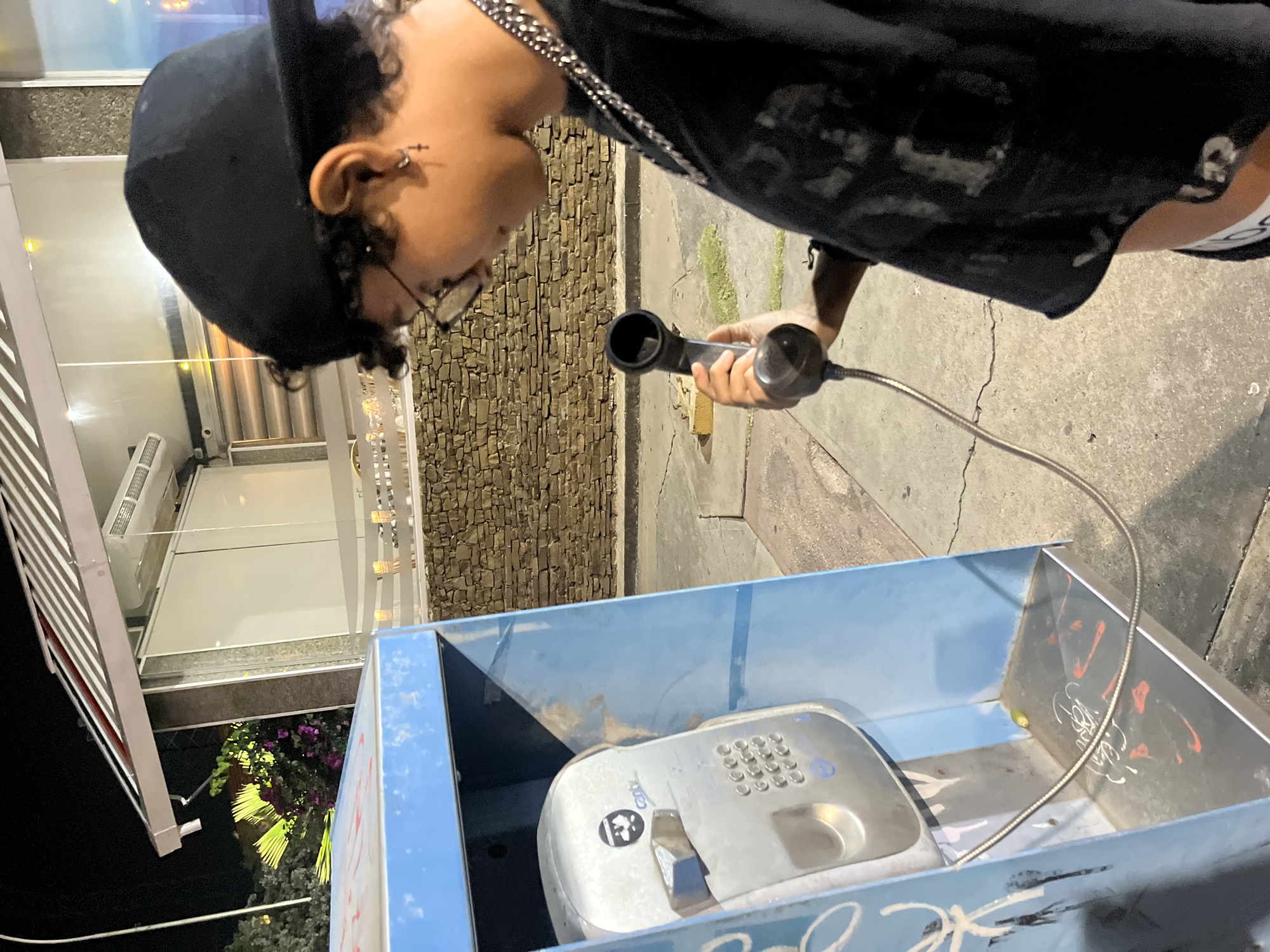 In the city of Pampatar, Venezuela, a young person wearing a black baseball cap, glasses, and a black t-shirt stands near a blue payphone booth. They are holding the handset of the payphone, which is an older model with a keypad and a slot for coins. The payphone is located on a sidewalk next to a building with a stone wall and a striped awning. The scene is illuminated by artificial light, suggesting it is nighttime. The payphone booth has graffiti on its interior. A green object, possibly a discarded item, is visible on the floor of the booth. The person is wearing a silver chain around their neck and a cross earring. The background shows a dimly lit street with some greenery and the facade of a building.