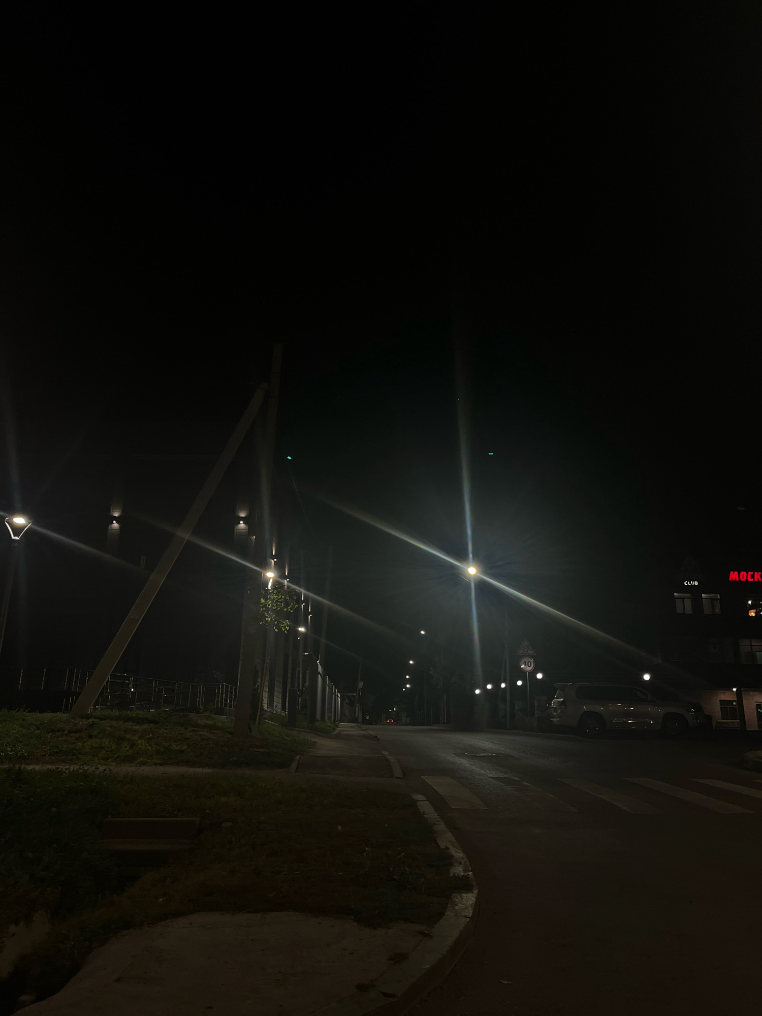 The image depicts a dark urban scene at night, with a street, sidewalk, and a grassy area on the left. Several streetlights are visible, casting a glow and creating lens flares. A building with illuminated signage, including "CLUB" and "MOCK," is present on the right side of the street. A white SUV is parked on the right side of the road, near a crosswalk. A speed limit sign indicating "40" is visible on a pole. The overall impression is of a quiet, dimly lit street in a city. The specific location in Almaty, Kazakhstan, is not discernible from the visual elements in the image.