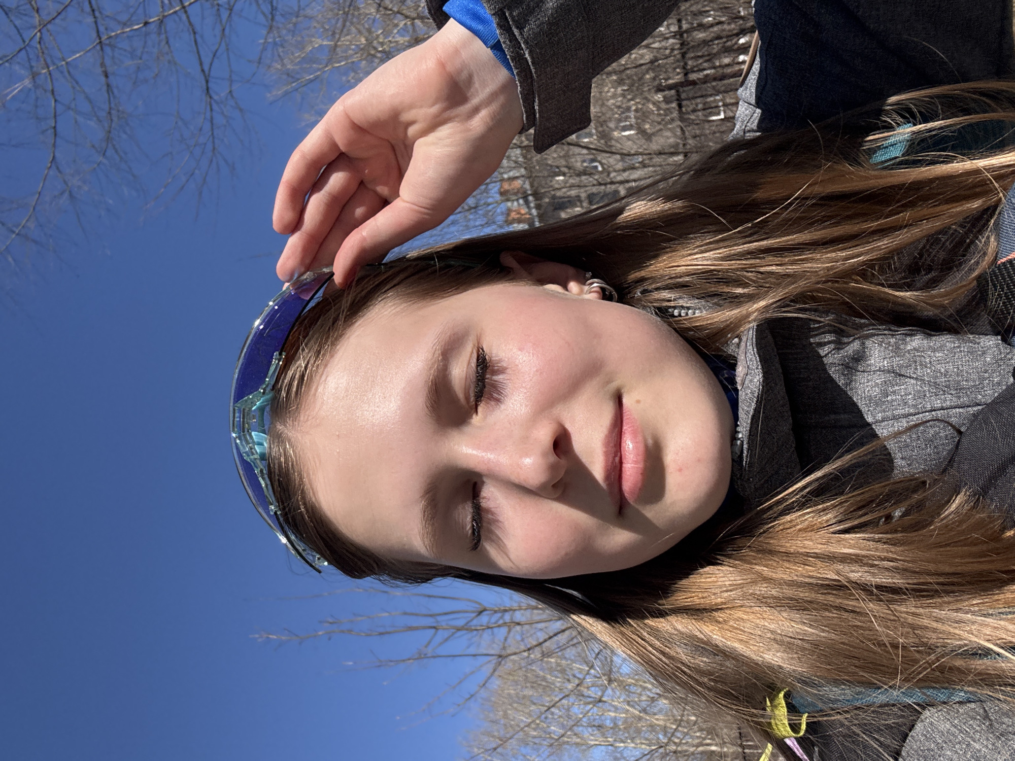 A medium shot of a person with fair skin and long brown hair, positioned outdoors against a bright blue sky partially obscured by bare tree branches. The person is wearing blue-tinted goggles pushed back on their head, held in place by their hand. They are also wearing a gray jacket with a blue shirt visible underneath. The setting appears to be a park or wooded area, but the location in Berezniki, Russia, cannot be confirmed from the image.