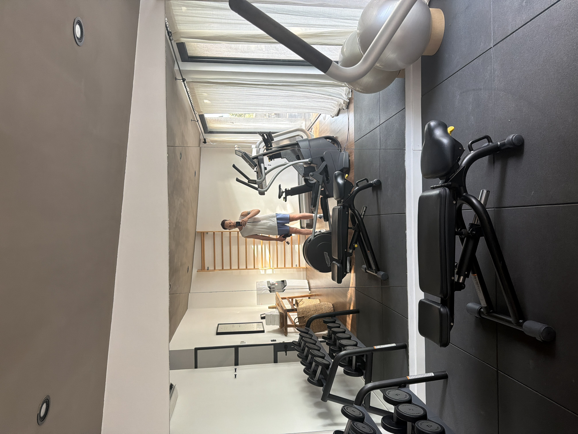 A man takes a mirror selfie in a modern home gym, reflecting a clean and well-equipped space. The gym features a variety of exercise equipment, including a weight bench in the foreground, a dumbbell rack to the left, and various cardio machines such as an elliptical and a stationary bike in the background. A large mirror covers most of the wall, reflecting the entire gym and the man. The environment appears to be a residential setting, possibly a home in Montuïri, Spain, given the natural light streaming through the sheer curtains in the background, suggesting a bright, sunny day. The man is wearing a white tank top and blue shorts, holding a phone to take the picture and a black water bottle in his other hand. The overall atmosphere is one of a private, dedicated space for fitness and wellness.