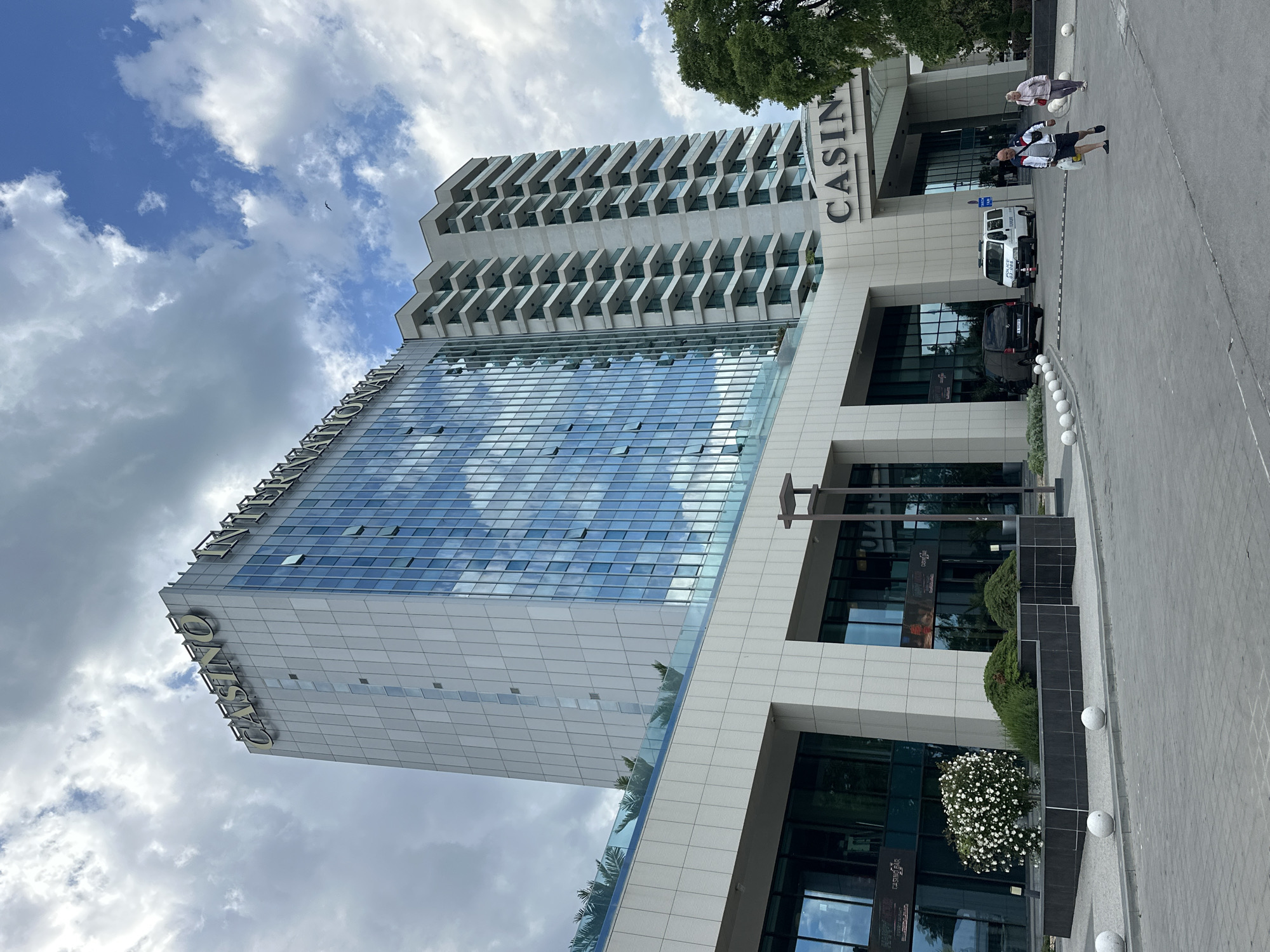 The image shows a tall, modern building in Varna, Bulgaria. The building is a casino, as indicated by the large "CASINO" sign on the side. It features a glass facade reflecting the cloudy sky. The building has a distinct architectural design with multiple floors and projecting balconies on the upper levels. In front of the building, there's a pedestrian area with planters, decorative white spheres, and parked vehicles, including a white van and a black car. Two people are walking on the sidewalk near the entrance of the casino. The angle of the image suggests it was taken from a low vantage point, looking upwards.