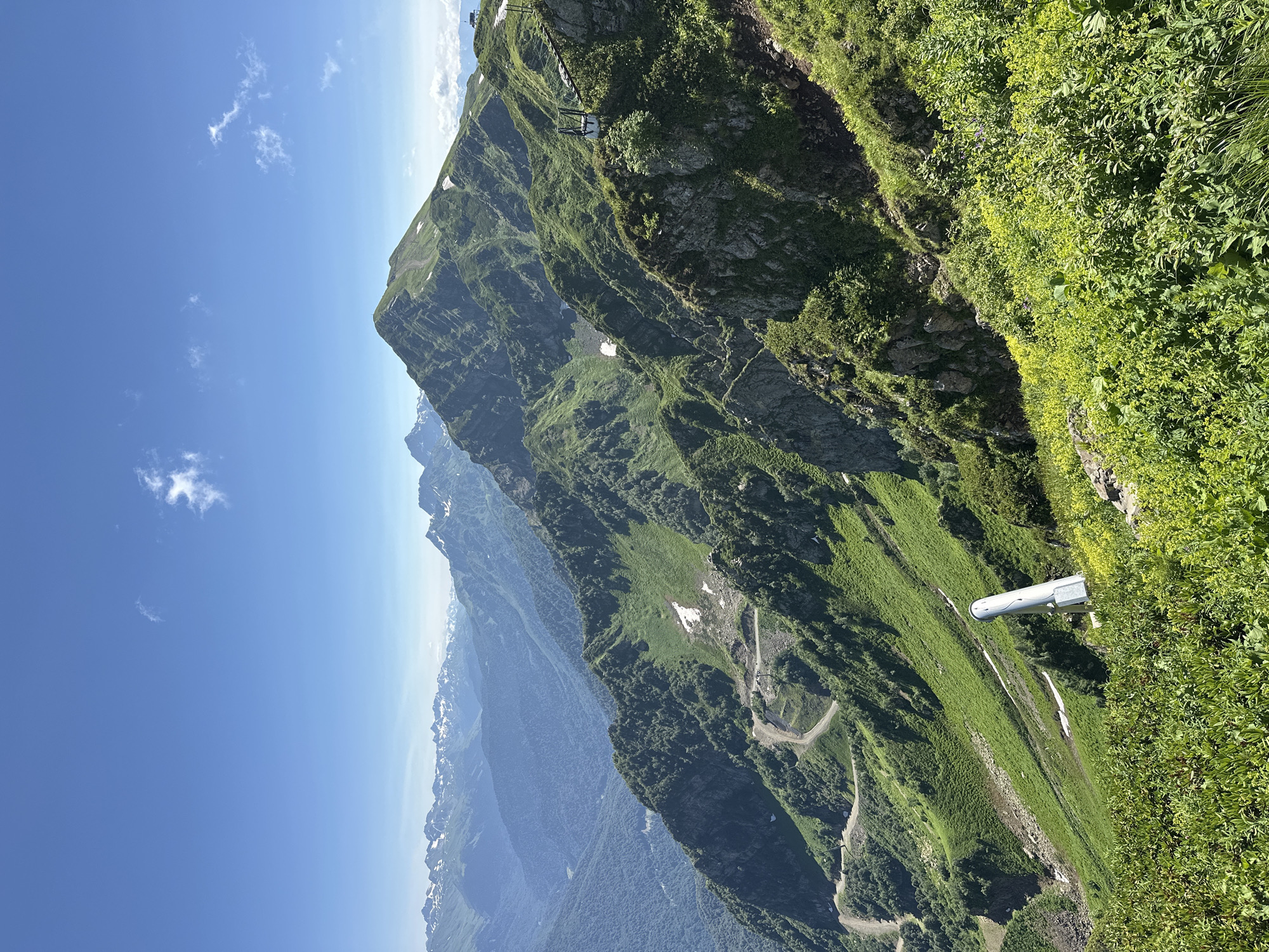 The image shows a vast mountainous landscape under a clear blue sky with scattered clouds. The foreground is filled with lush green vegetation, including flowering plants with yellow blossoms. In the middle ground, a steep, green mountainside is visible, with patches of snow. A winding dirt road or path is cut into the mountainside, and what appears to be a ski lift or gondola cableway structure is visible, along with a white cylindrical object protruding from the ground. Rocky outcrops and more vegetation cover the slopes. In the background, a series of rolling mountains recede into the distance, their peaks capped with snow. The overall impression is of a high-altitude, natural environment, likely a resort area, possibly in or near Estosadok, Russia, known for its mountain resorts.