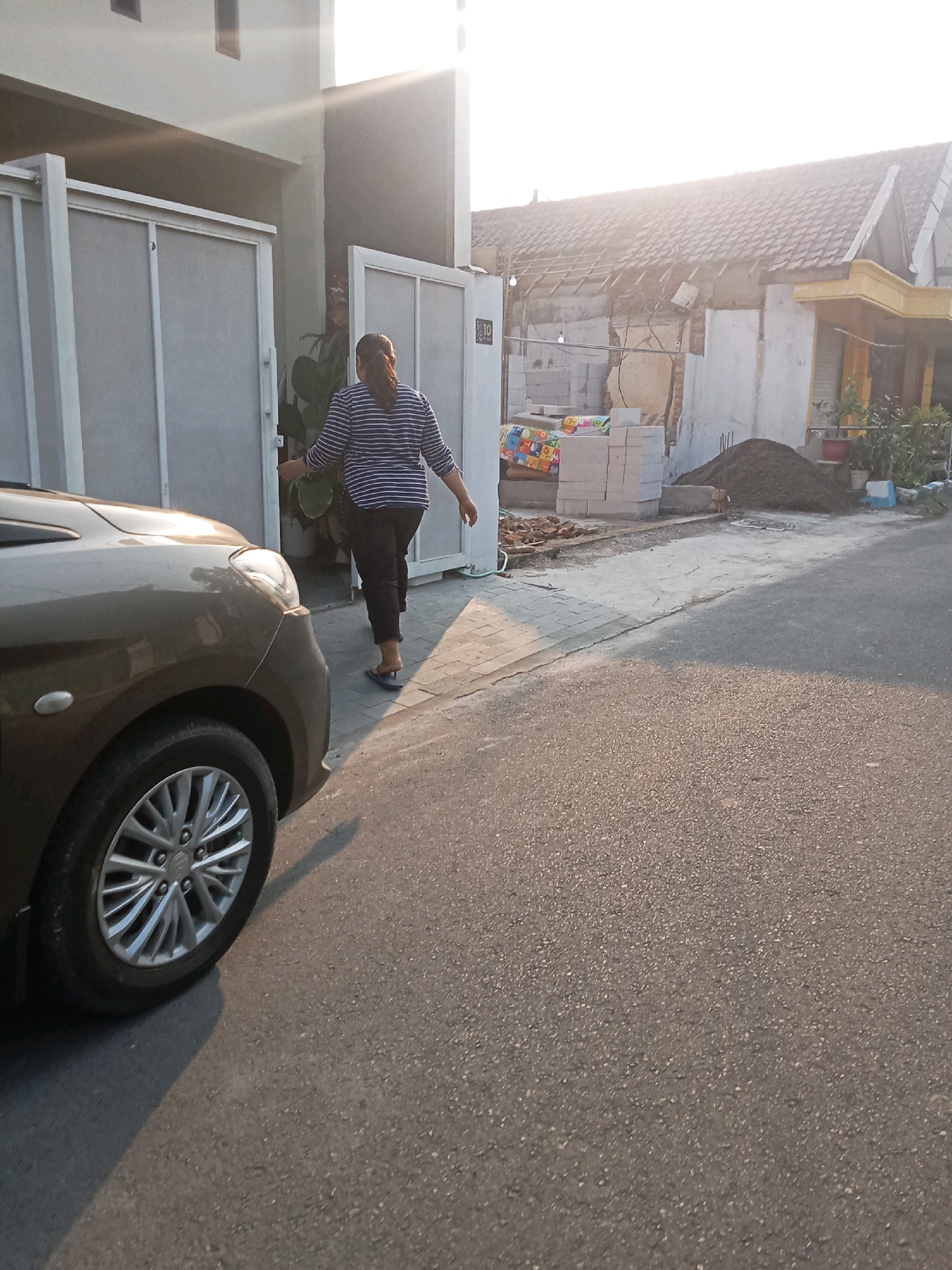 A woman in a striped shirt and black pants is walking towards a house entrance. She is exiting the frame to the right. A brown car is parked to the left of the frame, with its front tire visible. The scene is outdoors, with a paved road in the foreground and a house with construction materials in the background. The sun is shining brightly, casting shadows on the ground. The location is the city of Kaliwungu, Indonesia.
