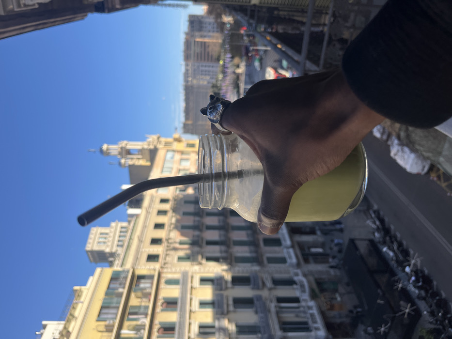 The image displays a first-person perspective from a balcony overlooking a city street in Barcelona, Spain. In the foreground, a hand with dark skin tone holds a clear glass mason jar containing a pale green liquid. A dark, bent reusable straw is inserted into the jar. A metallic ring, shaped like a wolf's head, is worn on the index finger. The background features several multi-story apartment buildings characteristic of traditional Catalan architecture, with balconies and facades in light yellow and beige tones. A prominent landmark, identifiable as the Casa Fuster, with its distinctive conical tower and dome, stands in the mid-ground, indicating a location near Passeig de Gràcia within the Eixample district. Below, a street is visible with numerous parked motorbikes and scooters. The sky is clear and bright blue, suggesting daylight with the sun casting long shadows from left to right. Blurred figures and vehicles populate the street below.