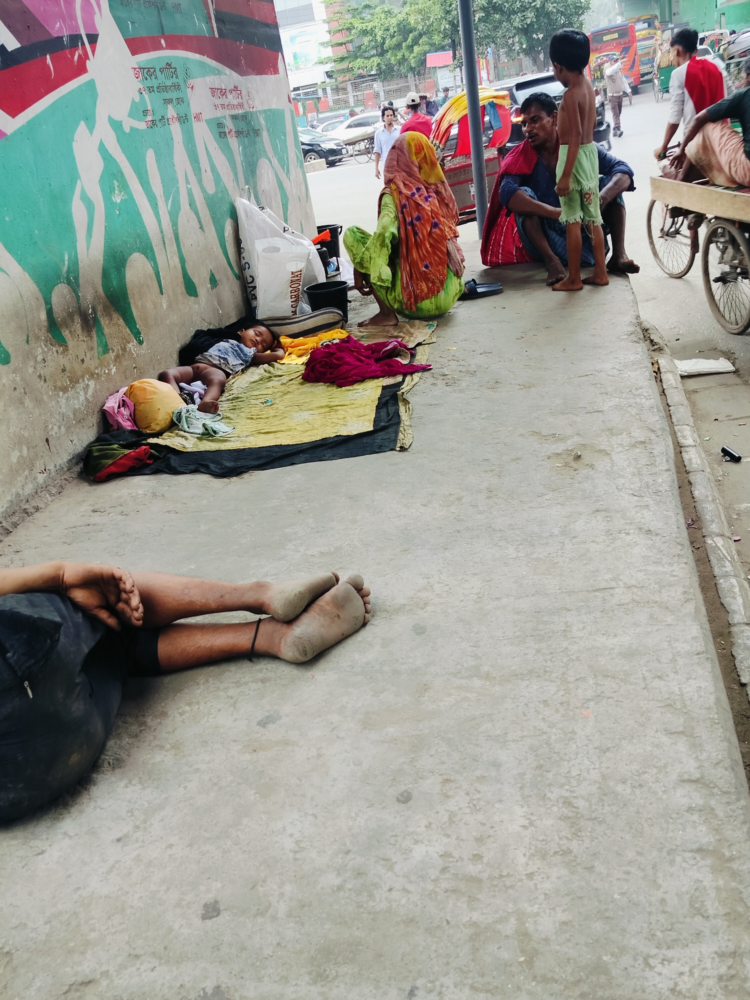 The image depicts a street scene in Dhaka, Bangladesh, focusing on a section of a sidewalk and its adjacent wall. A young child is asleep on a yellow blanket laid on the ground, near a black and white patterned mat. To the left of the sleeping child, a yellow pillow and folded clothing are visible.
Further down the sidewalk, an adult lies on their back, with their legs and feet prominently in the foreground. Their feet appear dusty.
In the background, several people are interacting. A man in a blue shirt is seated on the ground, facing a young boy who stands before him. The boy is shirtless and wearing green shorts. A woman in a bright green garment is also seated nearby. Behind them, a cart with two wheels is being pushed by a person in a red and white shirt. Other individuals and vehicles, including a colorful vehicle that appears to be a rickshaw, are present in the street, suggesting a bustling urban environment.
The wall behind the sleeping child is adorned with a large, colorful mural featuring stylized figures and text in Bengali. The sidewalk itself is made of concrete, showing signs of wear and tear. The overall scene captures a slice of daily life in a busy area of Dhaka, highlighting elements of poverty and community.