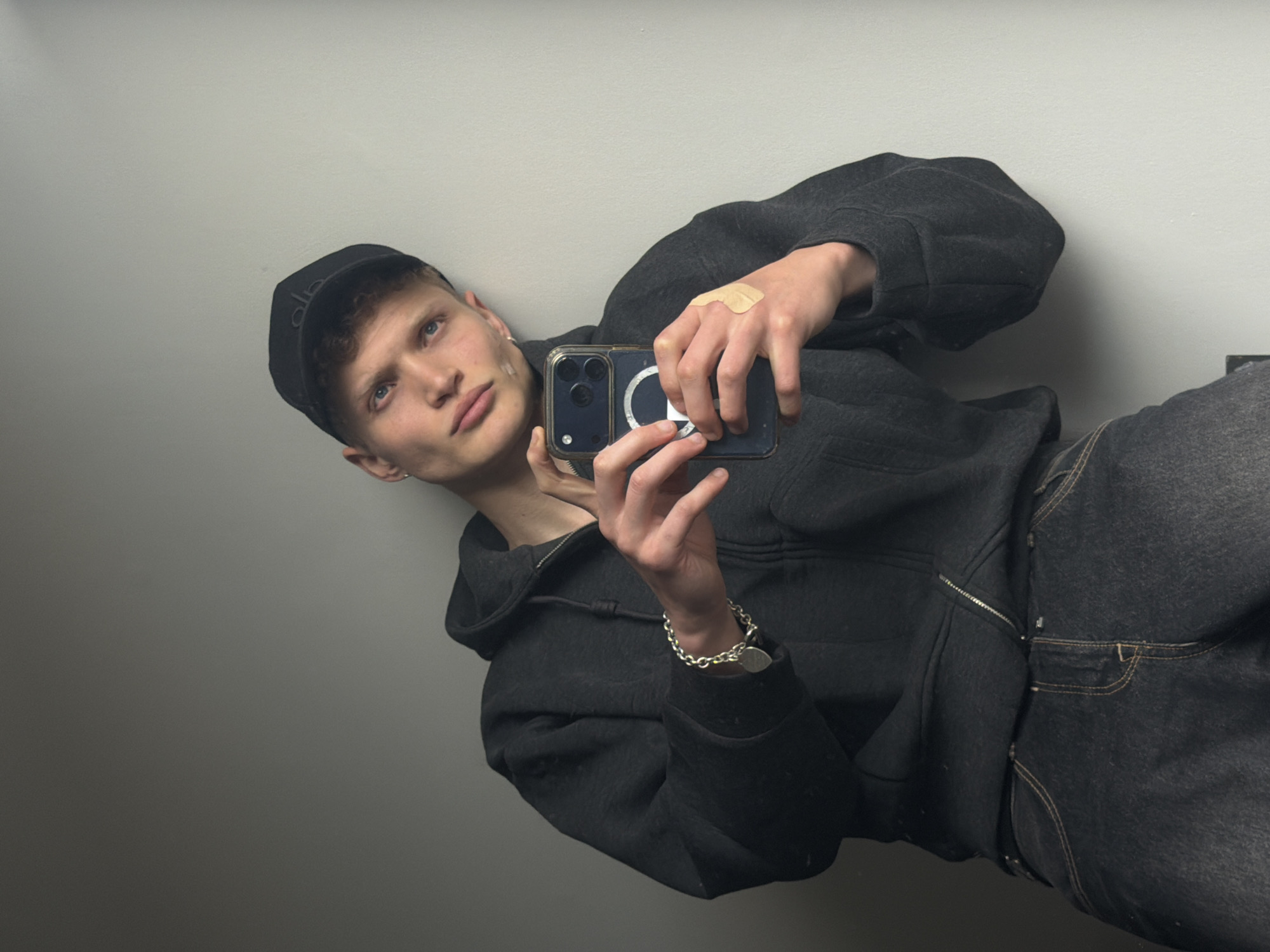 A young man with fair skin and short, light-colored hair is taking a selfie indoors against a plain, light gray wall. He is wearing a dark-colored hooded jacket with a zipper, dark jeans, and a black baseball cap. He is holding a smartphone with multiple camera lenses, likely an iPhone, in front of him, with his right hand near the top of the phone and his left hand near the bottom. A flesh-colored bandage is visible on the back of his right hand. He is also wearing a silver bracelet on his left wrist. Given the plain wall and lack of identifiable features, the location within London cannot be determined.