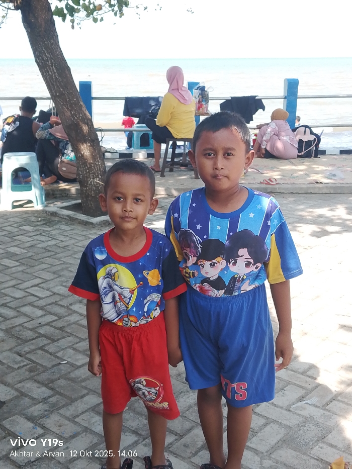 This image depicts a coastal area in Limbangan, Indonesia, under bright sunlight. In the foreground, two young boys stand side-by-side on a paved surface composed of grey rectangular pavers. The boy on the left, younger, wears a blue t-shirt with a red collar and sleeves, featuring an astronaut graphic, and red shorts. The boy on the right wears a blue t-shirt with yellow accents, displaying multiple anime-style male characters, and blue shorts with "BTS" printed in red on the left leg. Both boys have dark hair and face directly towards the camera.
In the midground, a brown tree trunk is positioned to the left. Behind the boys, a blue metal railing with white posts separates the paved area from a sandy beach. Beyond the railing, several individuals are visible. One person, wearing a yellow long-sleeved top and a pink hijab, is seated on a dark stool facing the water. Another person in light-colored attire is seated on the ground to their right. Dark garments are draped over the blue railing in two places.
The background shows a calm body of water, likely the sea, extending to the horizon under a bright, overcast sky. A narrow strip of sandy beach is visible between the railing and the water. The scene suggests a public recreational or leisure area by the coast.
