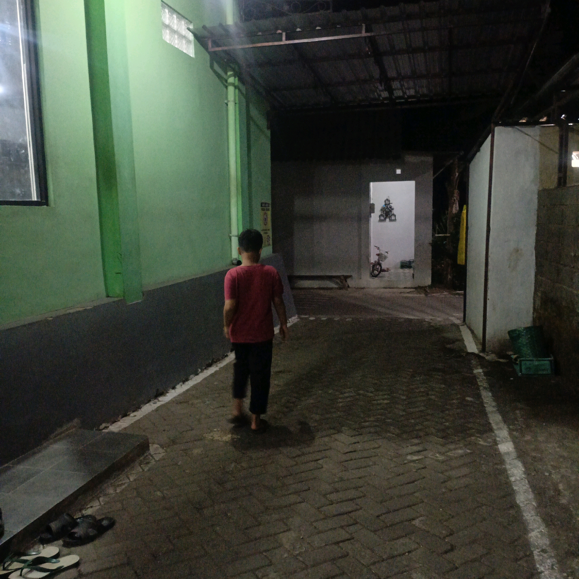 The image shows a boy walking away from the camera down a narrow, paved alleyway. He is wearing a red t-shirt and black pants. The alley is lined with green walls on the left and a more industrial-looking corrugated metal roof structure on the right. The ground is paved with dark bricks and is wet, reflecting the dim lighting. In the background, there are open doorways with glimpses of rooms inside. One doorway reveals a bicycle and some framed pictures on the wall. The scene appears to be an outdoor walkway in a residential or commercial area. The location is specified as Salatiga, Indonesia.