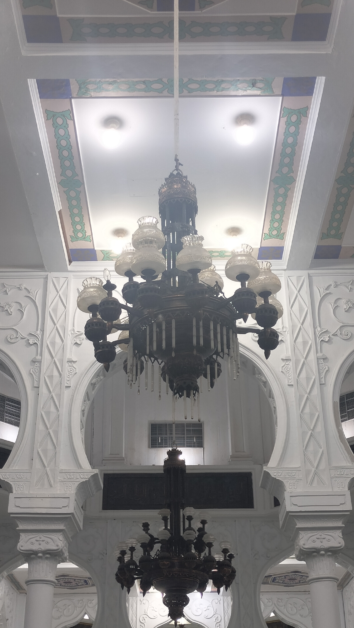 The image shows the interior of the Baiturrahman Grand Mosque in Banda Aceh, Indonesia. The focus is on two ornate chandeliers hanging from the ceiling. The larger chandelier, in the foreground, is dark metal with multiple arms holding glass lamp shades and decorative candle-like elements. The smaller chandelier, further in the background, is also dark metal with a similar design. The ceiling is adorned with decorative molding and intricate patterns in shades of white, blue, and green. The walls feature white plasterwork with arched doorways and decorative columns. The lighting is dim, with the chandeliers and ceiling lights providing illumination. The overall impression is one of grandeur and traditional Islamic architecture.