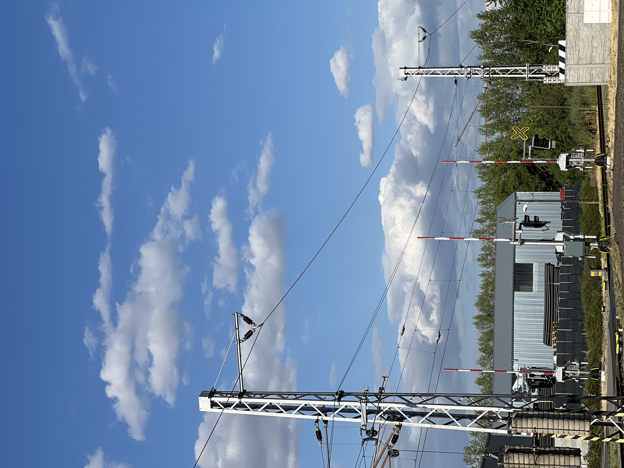 The image depicts a railway crossing with a clear blue sky filled with white, scattered clouds. In the foreground, two tall, white lattice metal poles support overhead electrical wires for the railway. These poles have various insulators and equipment attached. To the left of the image, a counterweight system is visible on one of the poles.
In the middle ground, there are multiple railway crossing gates with alternating red and white stripes. These gates are lowered, indicating the crossing is active or about to become active. Behind the gates, a corrugated metal building is visible, with stacks of black pipes or tubes on pallets in front of it. A dark fence runs along the perimeter of the building. To the right, another similar metal pole is present, supporting overhead wires. A yellow "X" shaped sign is visible on a pole, indicating a level crossing.
The environment is an outdoor setting, likely near a railway line in Újezd u Brna, Czechia. There are no people visible in the image. The overall scene is one of infrastructure related to a railway system, under a bright, partly cloudy sky.