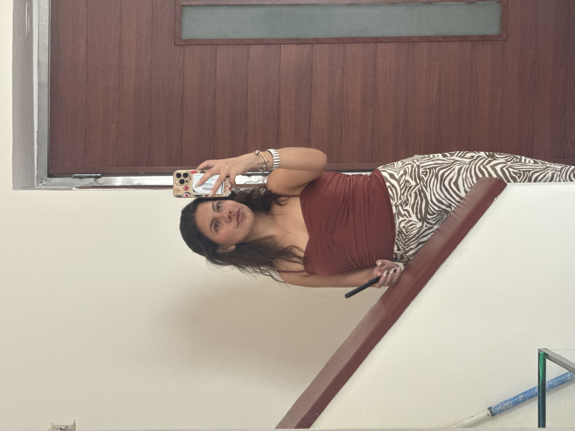 A young woman is taking a selfie on a set of stairs. She is wearing a rust-colored top and zebra-striped pants. She holds a black object in her left hand and a cell phone in her right. The background includes a brown door and a white wall. A metal railing is visible at the bottom right. The location is possibly in Heroica Puebla de Zaragoza, Mexico.