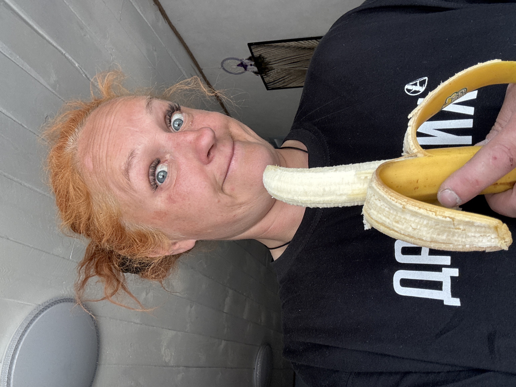A woman with red hair and blue eyes holds a peeled banana in front of her face. She wears a black t-shirt. The background consists of a gray-paneled wall, a round light fixture, and a framed picture hanging on the wall. There is no indication of the specific city of Chaikovsky, Russia in the image.