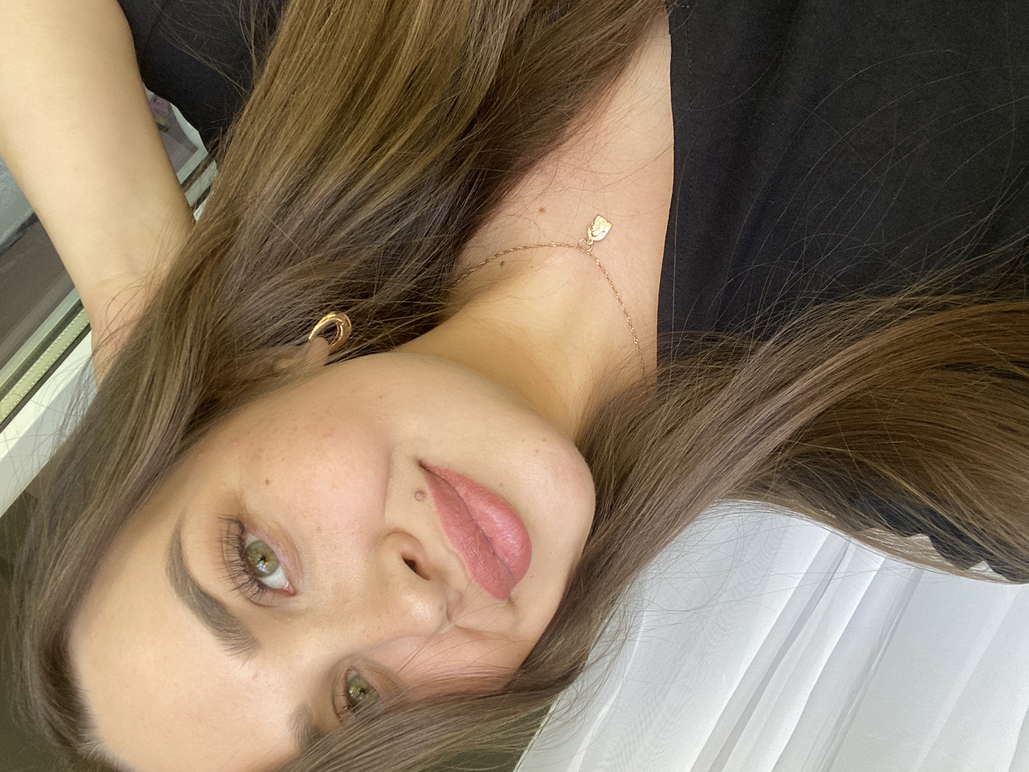 A young woman with green eyes and freckles lies down, tilting her head. She wears a black top and has brown hair, a gold necklace, and gold earrings. A white curtain is visible in the background. The location cannot be determined from the image.