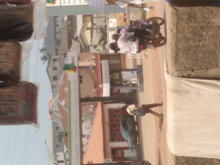 The scene depicts an active urban environment in Agege, Nigeria. In the immediate foreground, a light-colored, possibly off-white, fabric is draped over a low concrete wall, partially obscuring the bottom of the frame. Above, several retail items, likely hair products, are packaged in transparent plastic bags and suspended from an unseen overhead structure, slightly visible against the sky.
The midground features a dirt road with visible ruts and a depression. A male individual, wearing a light-colored polo shirt, dark trousers, and a blue and white patterned backpack, walks from right to left across this road. Further to the right, a motorcycle (okada) travels in the same direction, carrying three individuals: a driver, a primary passenger with dark hair, and a smaller, child-like passenger wearing patterned clothing seated behind the first. A dark-colored sedan is stationary on the left side of the road, adjacent to a building with an orange-brown roof.
In the background, a NNPC (Nigerian National Petroleum Corporation) gas station is prominent, featuring a white and green canopy structure with the corporation's logo visible. Several white and blue fuel pumps are positioned under the canopy. To the left of the gas station, a single-story building with a red roof and red-framed windows/doors is visible. Behind the gas station, a light-pink multi-story building with brown trim and exterior steps can be seen. The distant background consists of densely packed residential buildings of varying heights and colors, predominantly grey and white, with assorted roof types including corrugated metal and some flat roofs. Palm trees are interspersed among the structures, under a light blue, hazy sky.