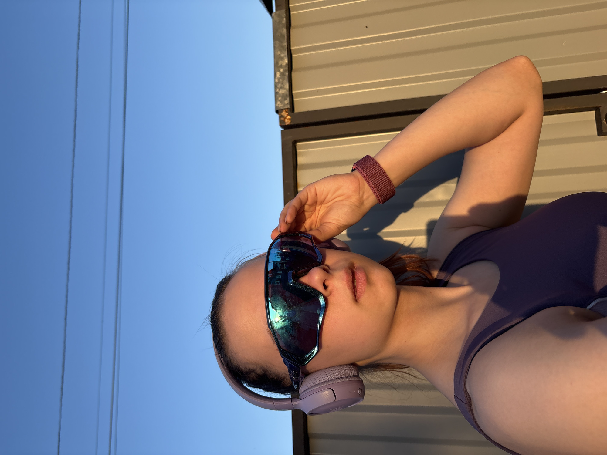 A woman wearing sunglasses and headphones stands outdoors in Прикамский, Russia. The woman is fair-skinned and wearing a purple top and a red wristband. She is holding her sunglasses with her right hand. In the background is a blue sky and metal siding. Overhead wires are visible in the upper left corner of the frame. The image is taken at an angle with the woman appearing to be leaning her head towards the camera.