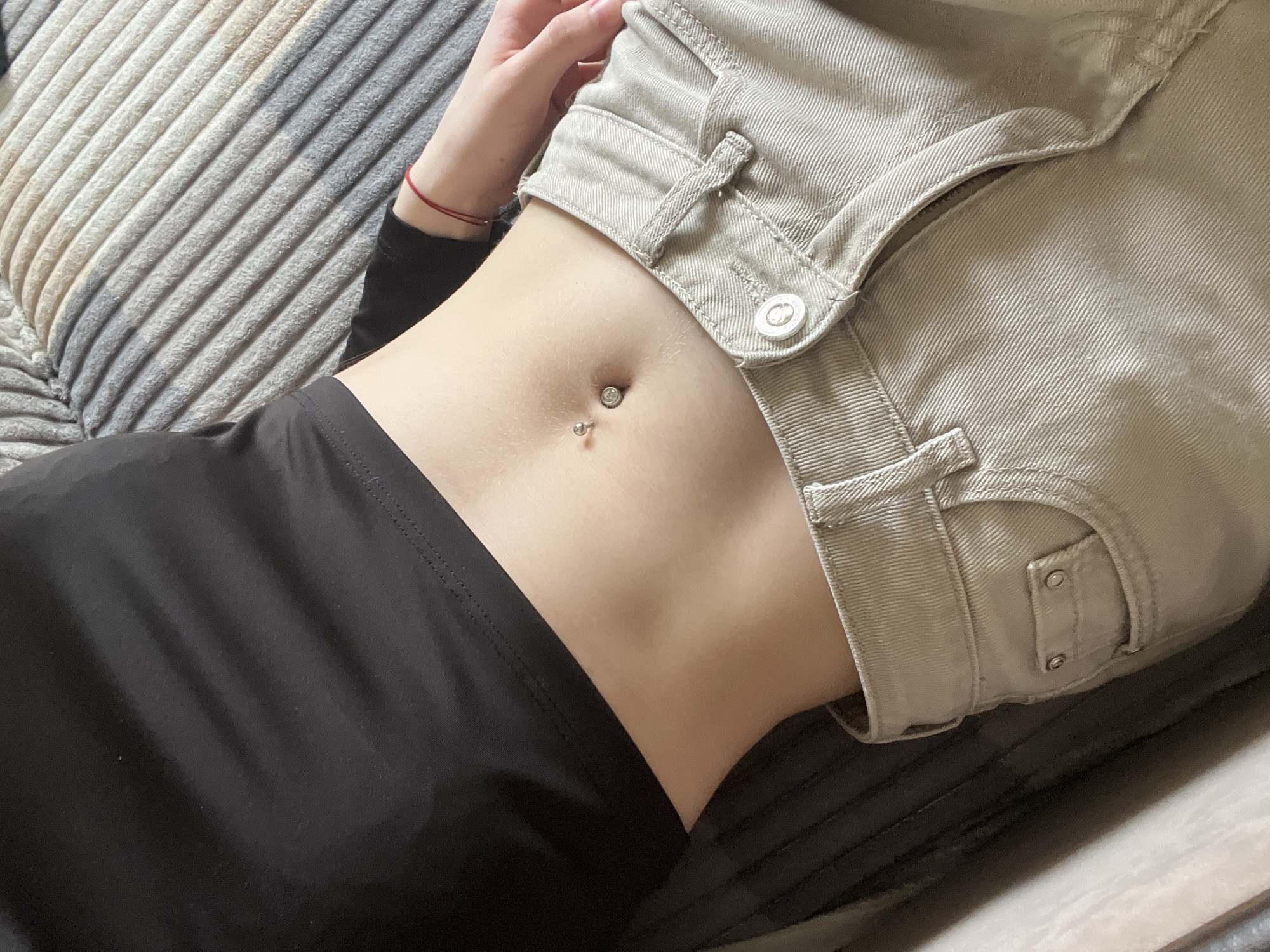 The image depicts a person lying on a grey, ridged surface, possibly a couch or bed. The person is wearing a black long-sleeved top and beige pants with the top button undone. A belly button piercing is visible. A red string bracelet is on their wrist. The location is unknown.