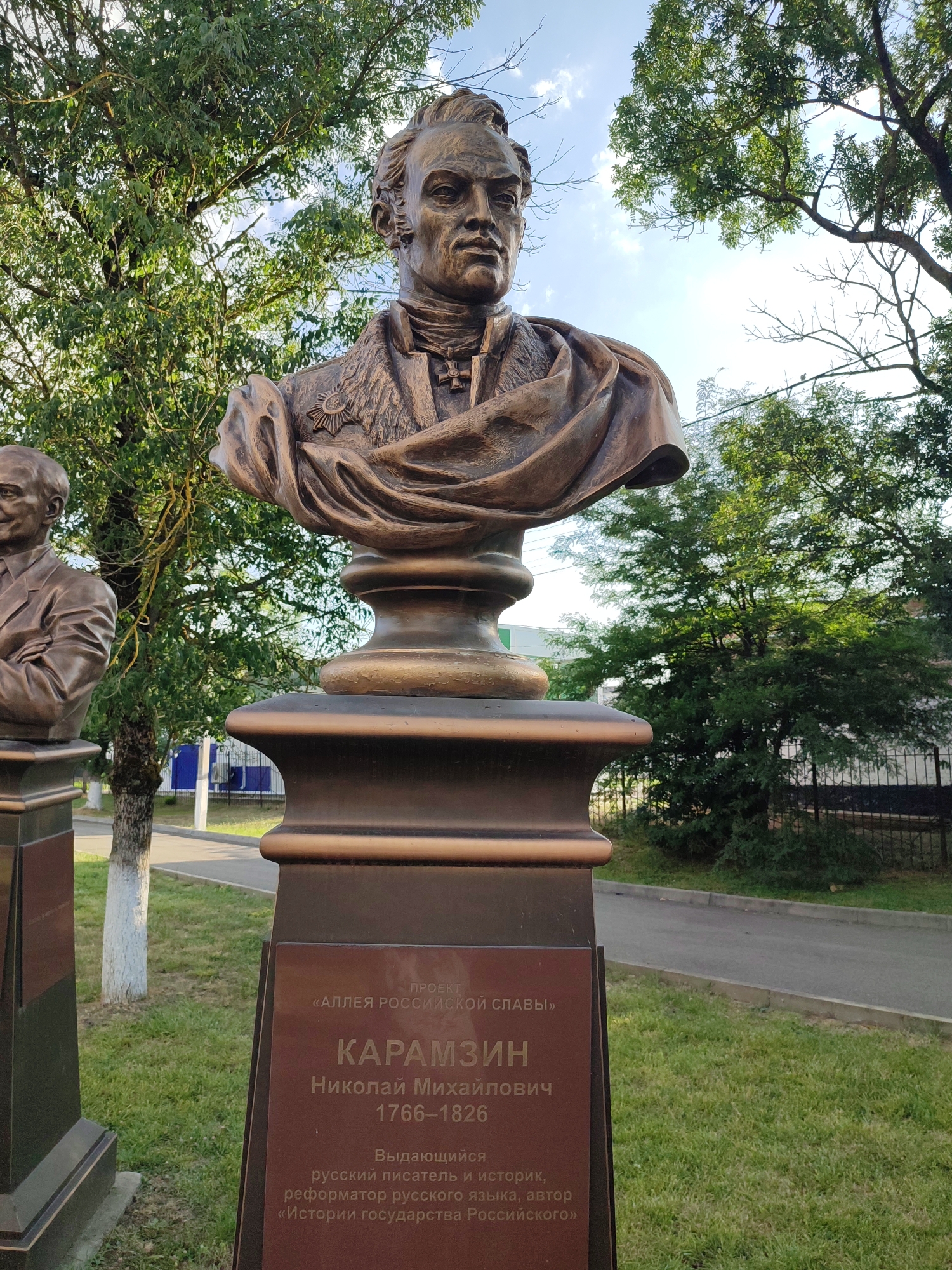 The image shows a bronze bust of Nikolai Mikhailovich Karamzin, a prominent Russian writer, historian, and reformer, located in the city of Abinsk, Russia. The bust is mounted on a pedestal with an inscription detailing his name, lifespan (1766-1826), and his contributions, including "History of the Russian State." The bust is positioned outdoors, with lush green trees and a blue sky with white clouds visible in the background. The lighting suggests it is daytime, with the sun casting soft shadows. To the left of Karamzin's bust, another bronze bust is partially visible, suggesting a collection or memorial of notable figures. The environment appears to be a public space, possibly a park or a square, with a paved walkway and grass in the foreground and mid-ground, and a fence and road visible in the distance.