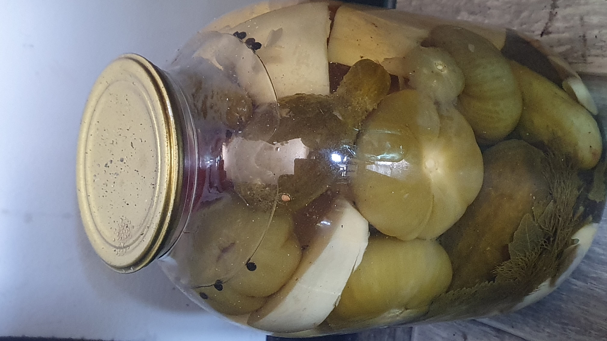The image shows a large glass jar filled with pickled green tomatoes and cucumbers, along with what appear to be slices of garlic and dill. The jar is sealed with a tarnished gold-colored metal lid. The contents are submerged in brine. The jar is placed on a wooden surface, likely a table or shelf, with a plain, light-colored background behind it. There are no people or animals visible in the image. The scene appears to be a domestic setting, possibly a kitchen or pantry, in the city of Ust-Labinsk, Russia.