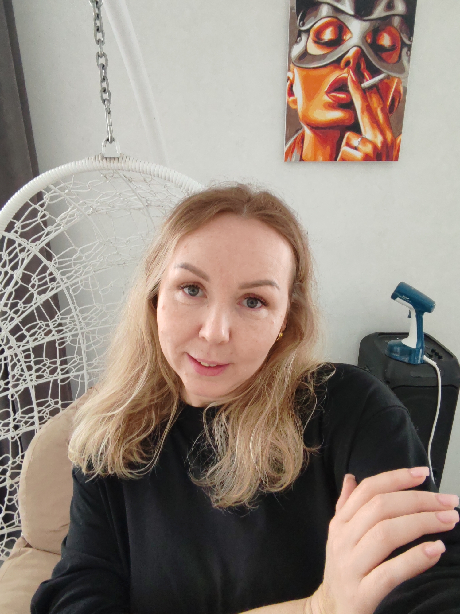 A fair-skinned woman with long, wavy blonde hair and light blue eyes is captured in a front-facing portrait. She is wearing a black long-sleeved top and is seated in a white, intricately woven hanging egg chair. Her left hand is partially visible, with light-colored fingernails. She looks directly at the camera with a calm, slightly smiling expression.
The setting appears to be indoors, likely a home in Chaikovsky, Russia. To the upper left, a metal chain supports the hanging chair, with a dark curtain visible in the background. A light-colored wall serves as the primary backdrop. To the upper right, a vibrant piece of wall art depicts a stylized woman wearing a silver mask, with prominent red lips and a finger pressed to them in a "shush" gesture, holding a lit cigarette. Below this artwork, on a black stand or surface, rests a small, blue and white handheld clothing steamer. The brand name "Jebat" is visible on the steamer. A white power cord extends from the steamer. The overall lighting is bright and even, suggesting daytime.
