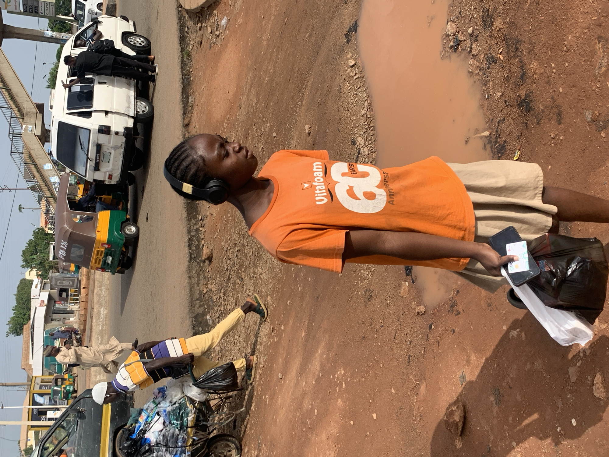 In Gura Top, Nigeria, a young person wearing black headphones and an orange t-shirt with a "Vitafom 60 Years Anniversary" graphic walks on a dirt road. They are holding a black plastic bag, a white plastic bag, and a smartphone. The ground is reddish-brown dirt, with a large puddle of muddy water to the right of the person. In the background, a white van is parked, with people standing around it. Several auto-rickshaws, also known as Keke Napeps, are visible, along with other vehicles and buildings. An overhead pedestrian bridge crosses the road in the distance. The scene depicts a busy street with various modes of transportation and activity typical of a Nigerian urban environment.